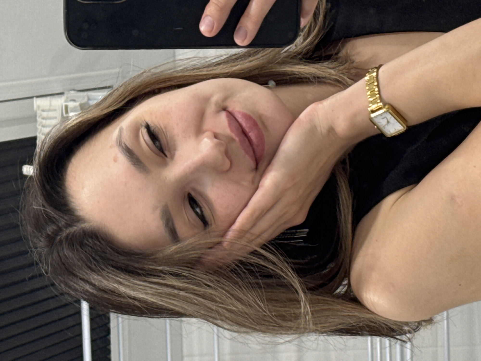 The image shows a woman indoors. She is holding a smartphone to take a selfie. She is resting her face on her hand. She is wearing a gold watch. Her hair is long, light brown, with darker roots. She is wearing a black top. The background is a white wall with a metal grid. The location and city are not discernible from the image.