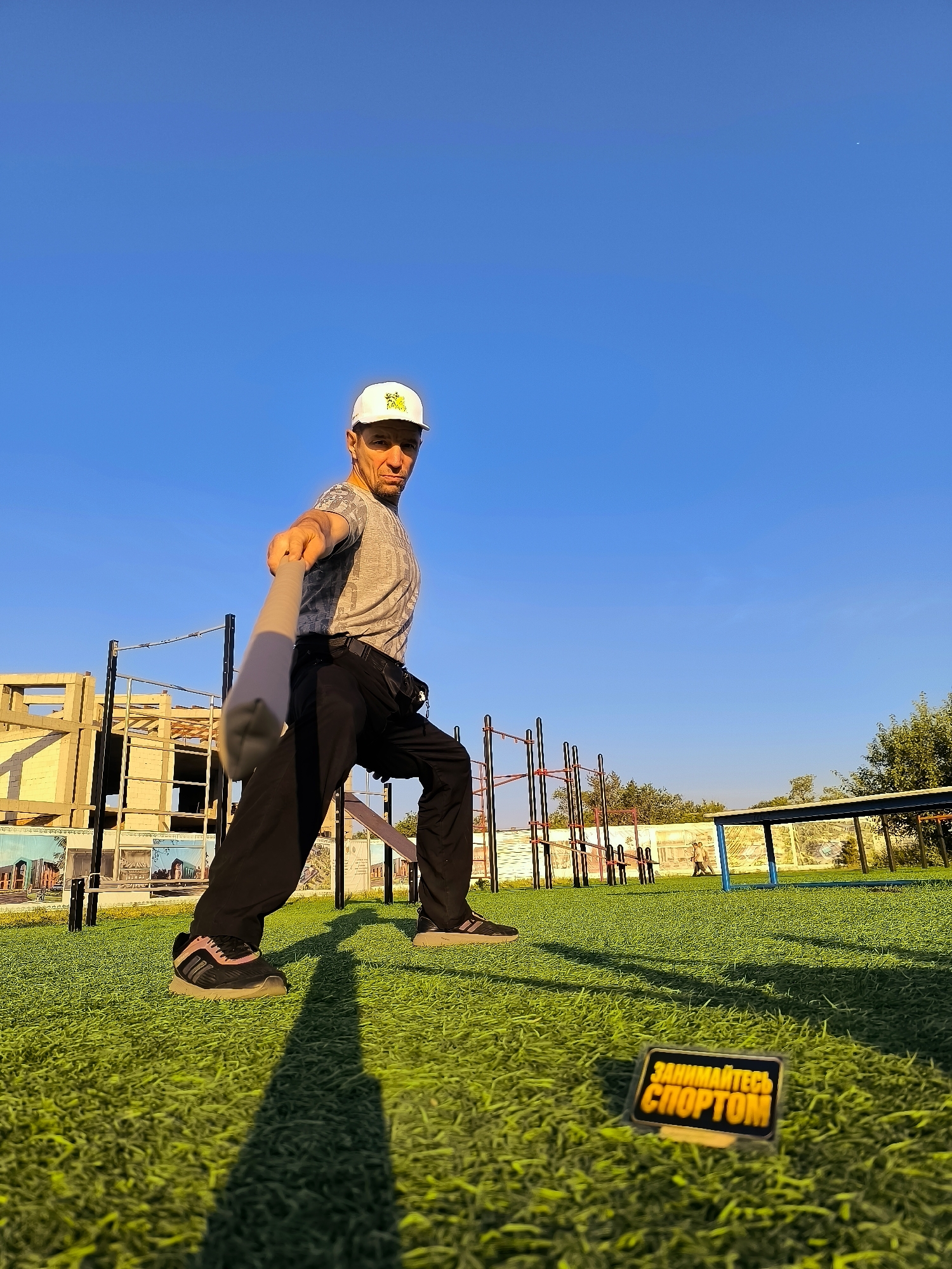 A man wearing a grey t-shirt, black pants, and a white baseball cap stands on a bright green artificial turf field in a park. He is holding a grey object, possibly a baseball bat or stick, horizontally in front of him with his right arm extended. His left leg is forward, and his right leg is bent, suggesting a dynamic pose or action. Behind him, there are various exercise bars and a partially constructed building with scaffolding. A small black and yellow sign with Russian text, "ЗАНИМАЙТЕСЬ СПОРТОМ," meaning "DO SPORTS," is visible in the foreground on the grass. The sky is a clear, vibrant blue. The overall scene depicts an outdoor sports or training area.