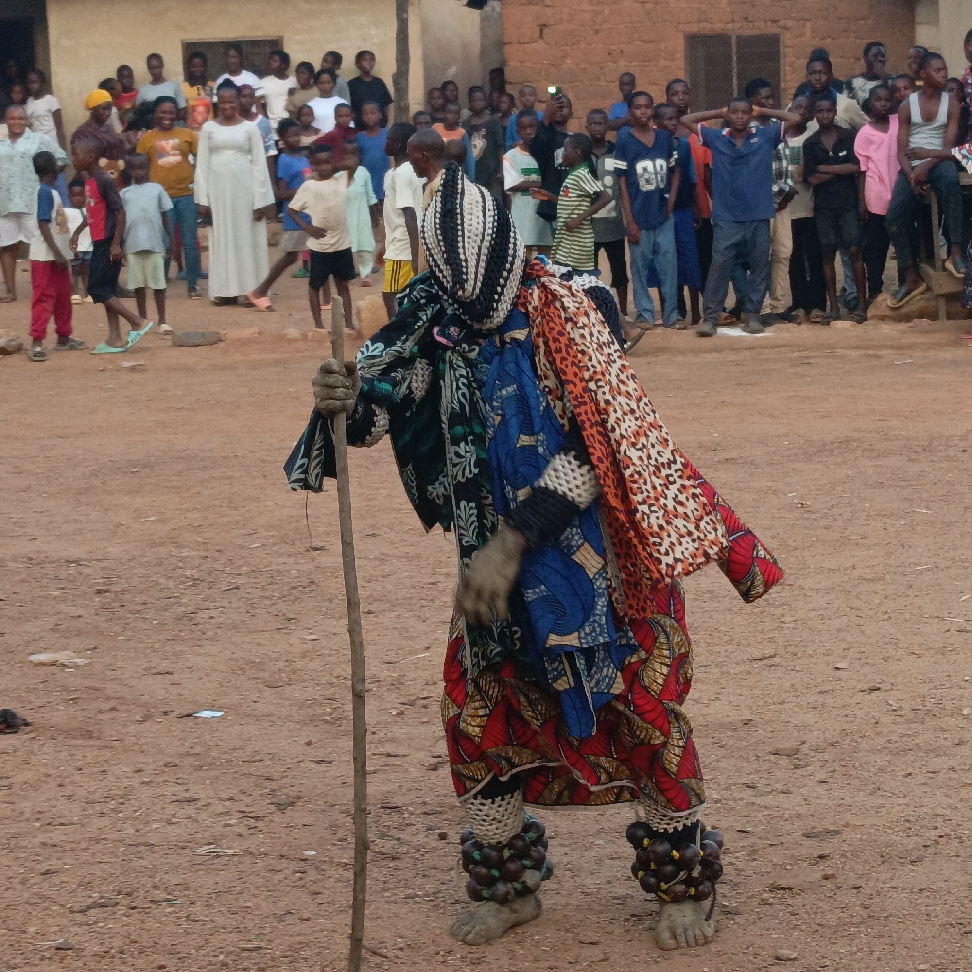 In Akwanga, Nigeria, a person in ceremonial attire stands in a dirt clearing before a crowd. The person is wearing a multi-colored, patterned garment. Their head is covered with a black and white patterned mask. They hold a wooden staff in their right hand. The bare feet are adorned with anklets. A crowd of men, women, and children stands in the background. Buildings are visible in the distance.