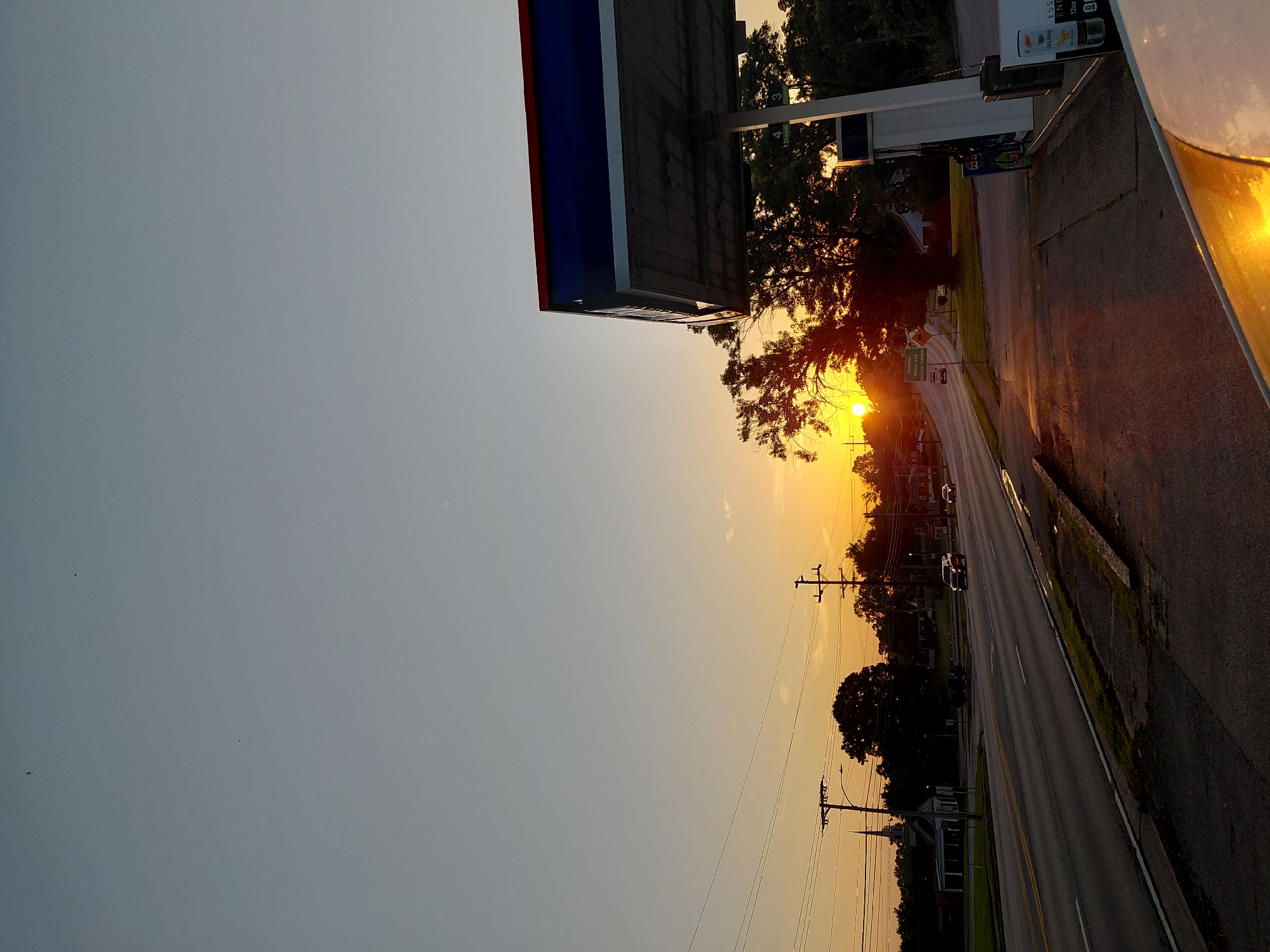 The image depicts a sunset scene at a gas station in Frankfort, United States of America. The sun is low on the horizon, casting a warm glow and long shadows. The sky is a gradient of pale blue to orange. A gas station canopy, with blue and red trim, is prominently featured in the foreground, partially obscuring the view. To the left of the canopy, a road with traffic is visible, with cars driving and a church steeple in the distance. Trees and power lines line the road. The ground in front of the gas station pumps is paved, and a portion of a white vehicle is visible in the lower right corner.