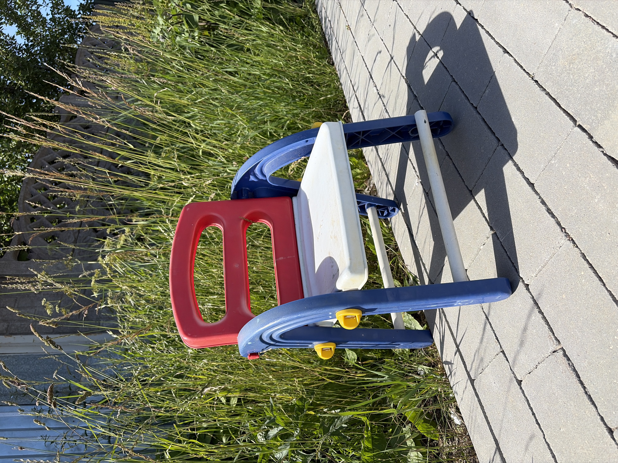 A red and blue plastic child's chair sits on a paved walkway in front of a wall and tall grass. The chair has a red back and seat with blue armrests and legs. The seat is slightly dirty. The walkway is made of gray rectangular pavers. The grass is green and overgrown, reaching up to the top of the chair. A shadow of the chair is cast on the walkway. The background is blurred, with hints of a fence and trees. The location is Unknown, Unknown.