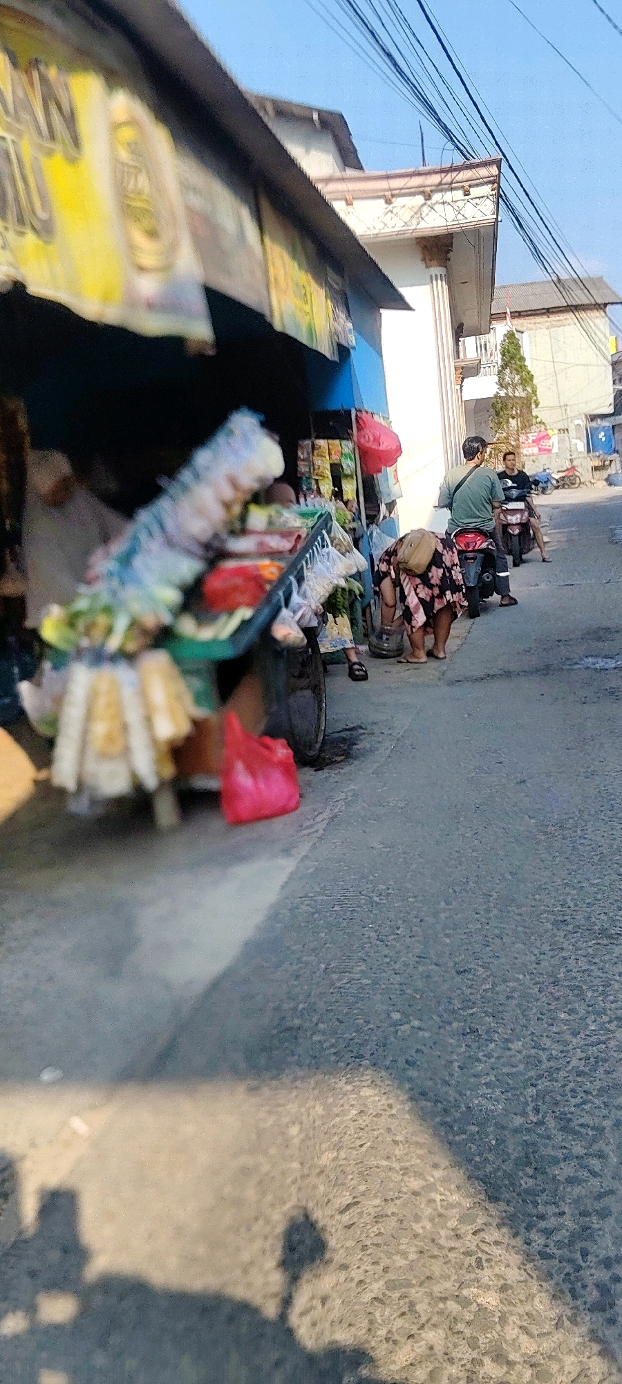 The image depicts a street scene in Mekarmukti, Indonesia, on a sunny day. In the foreground, a shadowy silhouette of a person is cast on the asphalt road. To the left, a market stall is visible, with various goods displayed on a cart. The stall has a blue awning and a yellow banner with text that is difficult to read due to blurriness. Several items, including plastic-wrapped goods and bags, are arranged on the cart.
Further down the street, a woman wearing a floral dress is bending down, possibly attending to something on the ground. She has a light brown bag slung over her shoulder. To her right, a man in a green shirt is seated on a motorbike. Another man is on a motorbike further down the street. The street is paved and appears to be in a built-up area, with buildings visible in the background. Electrical wires are strung across the sky. A red plastic bag lies on the ground near the market stall. The overall impression is of a bustling street in a residential or commercial area.