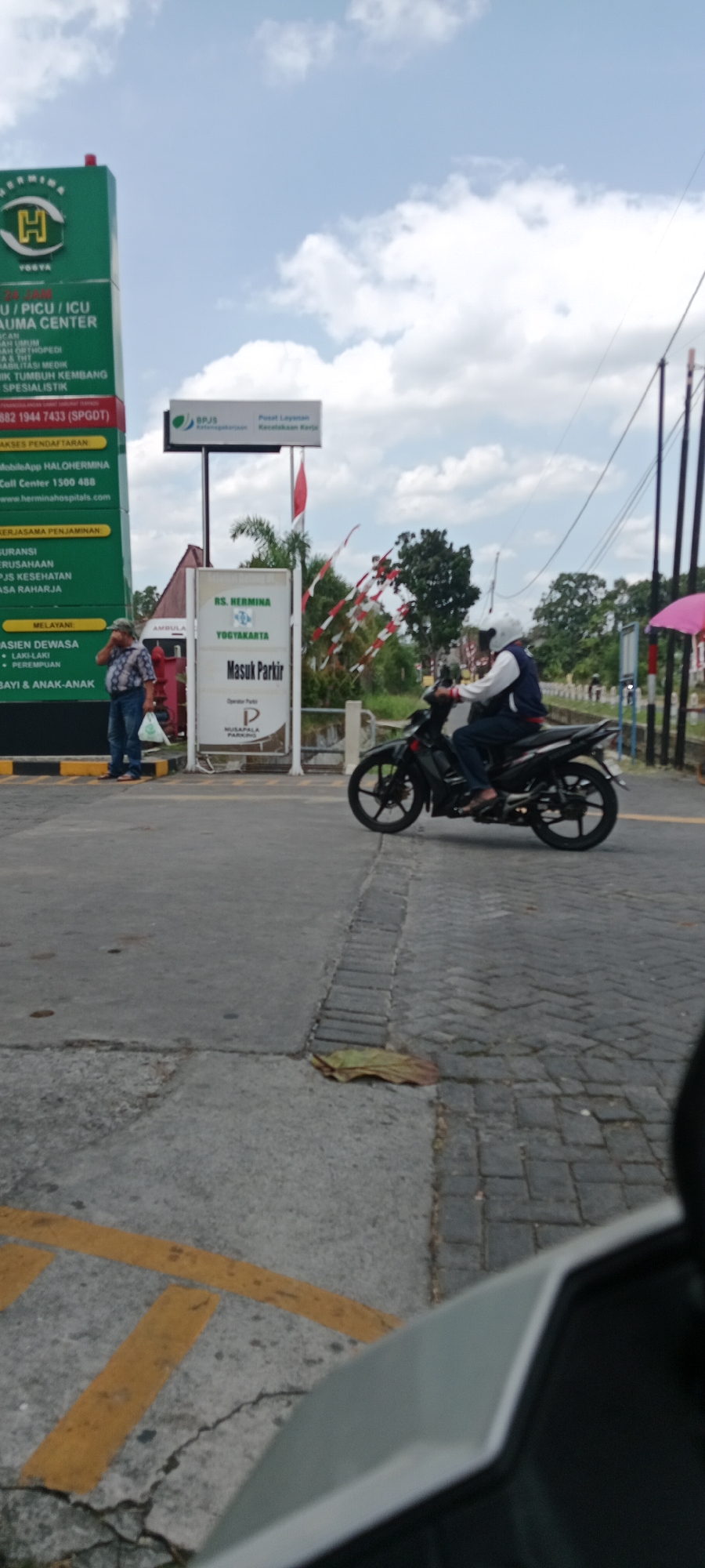 The image depicts an outdoor scene near a hospital entrance in Maguwoharjo, Indonesia. A tall green sign for "RS. HERMINA YOGYAKARTA" with a list of services and contact information is prominently displayed on the left. Adjacent to it, a white sign indicates "MASUK PARKIR" (Enter Parking).
A man in a checkered shirt and jeans is standing near the green sign, holding a plastic bag. A motorcycle with two occupants, both wearing helmets, is in the process of entering the parking area, with the motorcycle on the right side of the frame. The road surface is paved with concrete and some cobblestone sections.
The environment includes a blue sky with scattered clouds, trees in the background, and a red and white flag attached to a pole. Utility poles and wires are also visible. The scene suggests a daytime setting with moderate cloud cover.