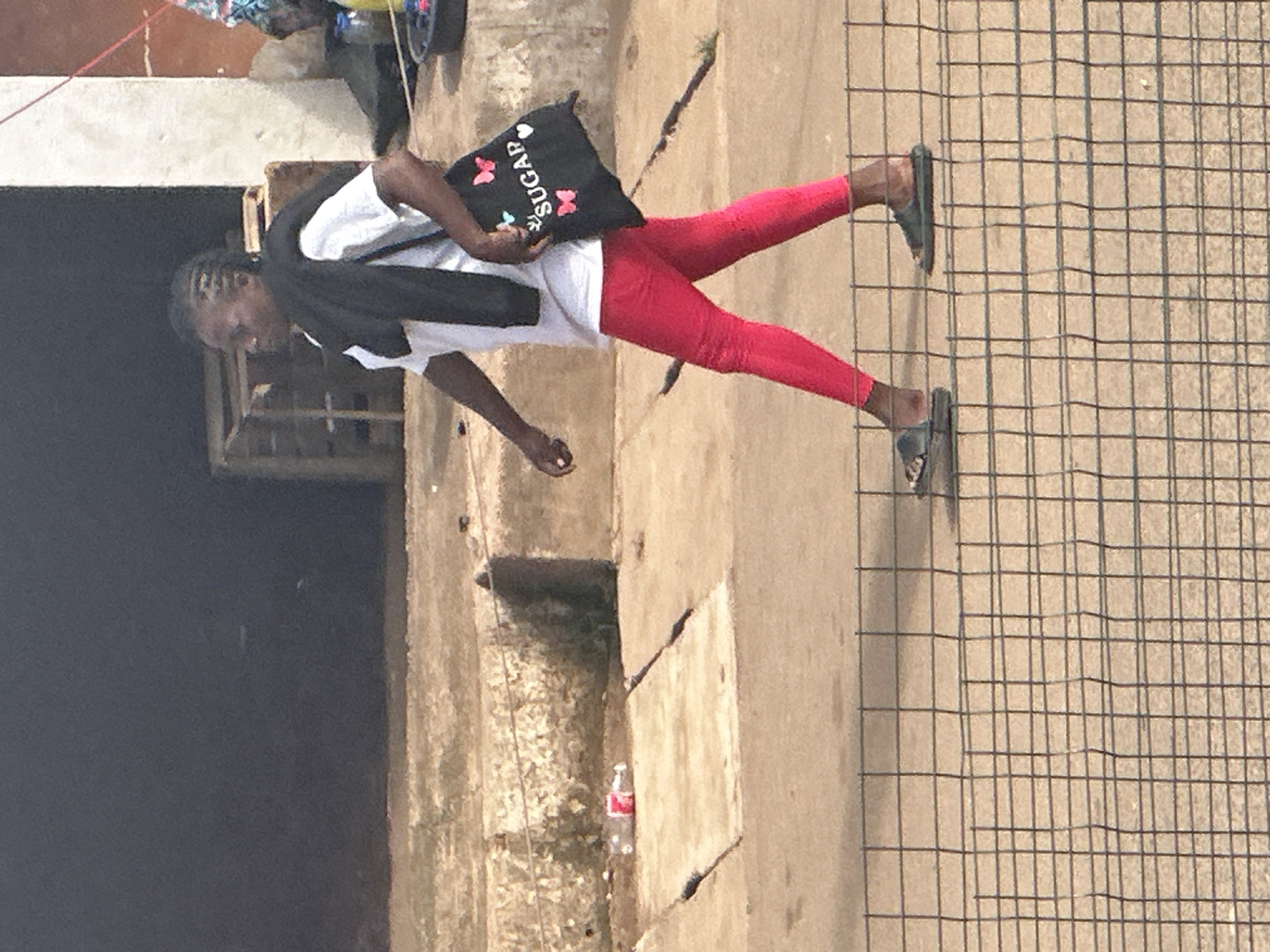 In Bukuru, Nigeria, a woman with braided hair, wearing a white t-shirt, black scarf draped over her shoulders, and bright pink leggings, walks past a wire fence. She carries a black tote bag adorned with butterflies and the word "SUGAR" in white lettering. The background reveals a coarse, sandy ground and what appears to be a rough concrete structure, possibly part of a building or walkway, with a discarded plastic bottle lying on it. The lighting suggests an outdoor, daytime setting.