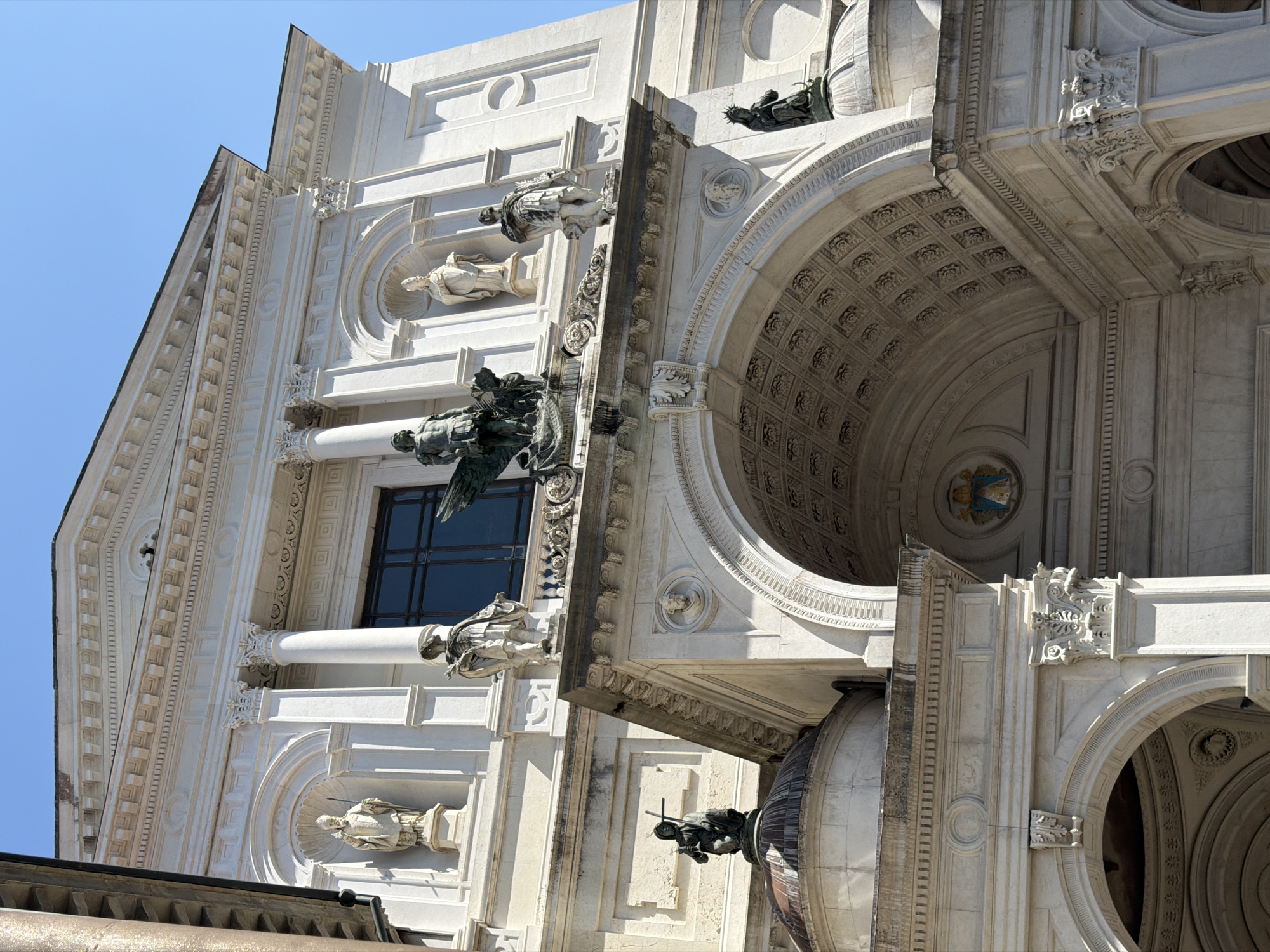 The image shows a detailed view of the facade of the Church of San Lorenzo Martire in Mozzo, Italy. The church's white stone exterior is adorned with classical architectural elements, including columns, pediments, and statues. At the center of the facade is a large arched entrance, above which sits a sculptural group featuring a winged figure. Niches along the upper facade house statues of various figures, likely saints or religious icons. The clear blue sky suggests it is daytime. The building exhibits intricate carvings and decorative friezes.