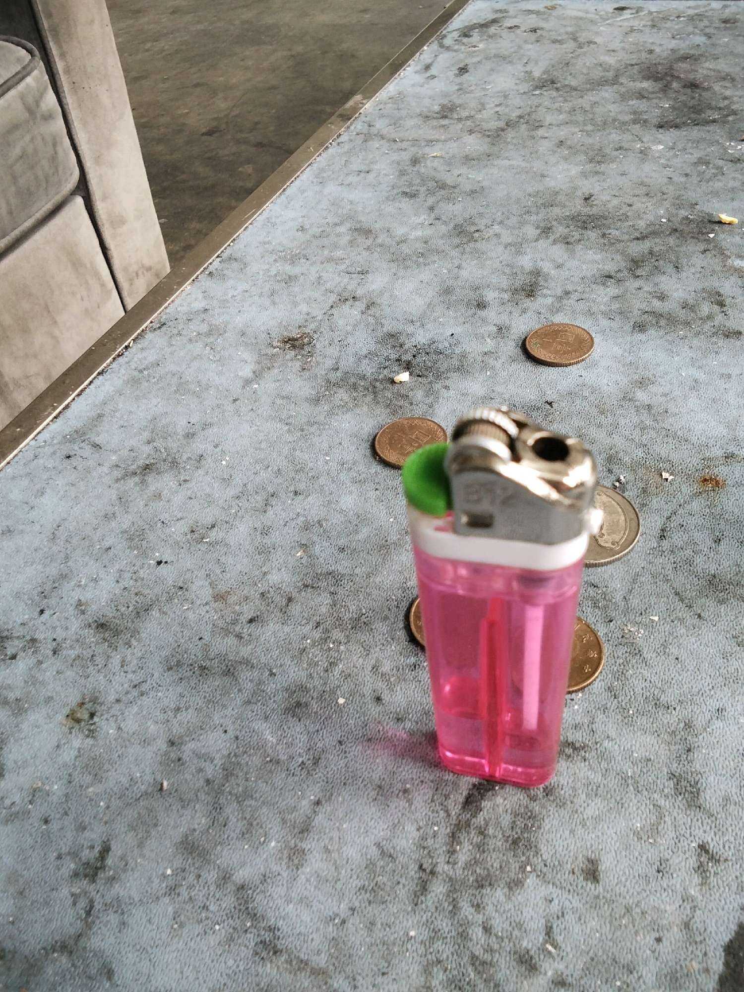 A pink disposable lighter sits on a dirty, textured blue surface, possibly a table. The lighter is surrounded by several gold-colored coins, some of which are partially visible. The surface has numerous dark smudges and some lighter speckles, suggesting it is not clean. In the background, a portion of a light-colored couch or chair is visible on the left, and a metallic strip runs along the top edge of the blue surface. The scene appears to be indoors, in a location that is possibly dusty or neglected. There are no people visible in the image.