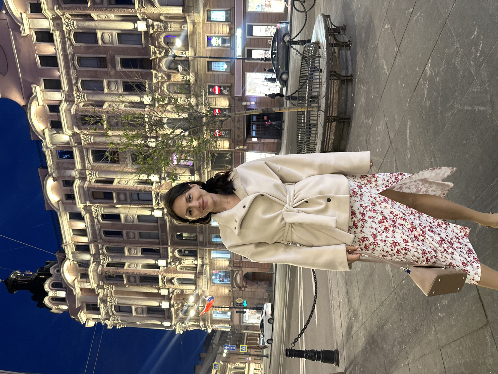The image depicts a woman standing on a sidewalk in Saint Petersburg, Russia. The woman is wearing a white coat over a floral dress and carrying a small purse. The background features an ornate, illuminated building, likely a historic structure typical of the city. Streetlights and other city infrastructure are visible, indicating a nighttime setting. There's also a bench in the scene.