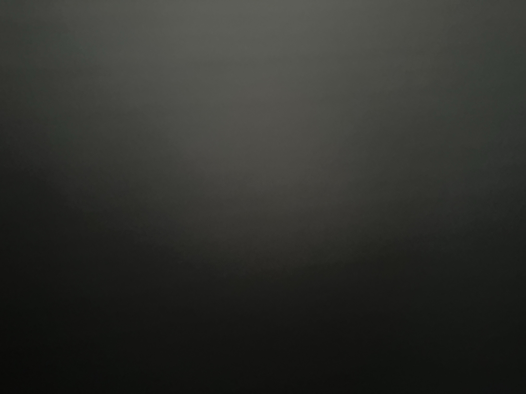 This is a low-light, abstract, and out-of-focus image of a dark, textured surface, likely a wall or fabric. The lighting is dim, creating a gradient from a slightly lighter, diffused gray in the upper portion of the image to a deep, almost black tone at the bottom. There are subtle variations in shade and texture throughout, suggesting a rough or uneven surface. The overall impression is one of darkness and obscurity, with no distinct subjects or activities visible. The lack of detail makes it impossible to determine the time of day, weather, location cues, or emotions. There is no visible text in the image.