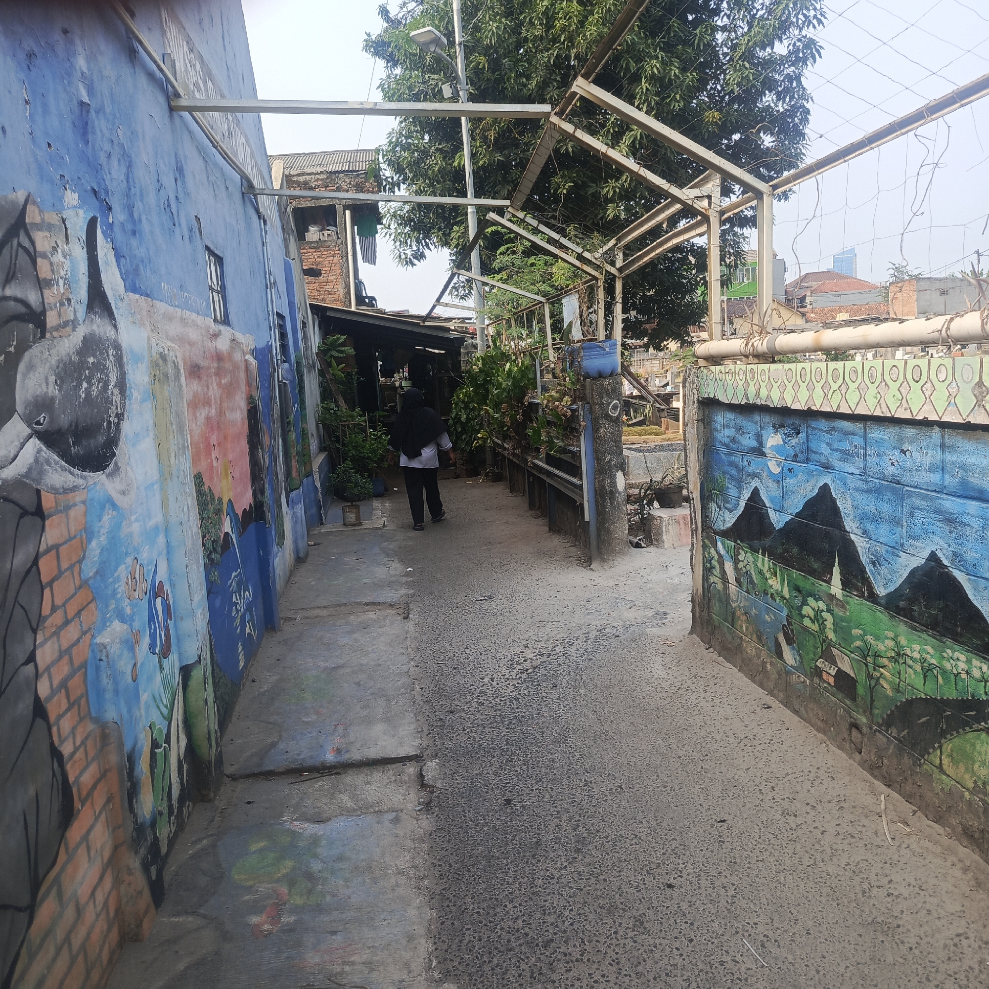 The image depicts a narrow alleyway in South Jakarta, Indonesia, characterized by vibrant murals adorning the walls. On the left, a blue wall features a large mural, partially faded, with depictions of what appears to be a bird's head, brickwork, and marine life such as fish. The alleyway itself is paved with asphalt in the center, transitioning to concrete on the left side. A person wearing a black hijab and a light-colored shirt over black pants walks away from the camera down the alley. To the right, a wall is painted with a landscape mural showing dark mountain peaks against a blue sky with a moon, and a green foreground with small houses and trees. A metal structure with a wire mesh runs overhead, suggesting a possible canopy or support system. Vegetation, including potted plants and trees, is visible along the sides of the alley and in the background, interspersed with buildings and structures typical of an urban environment. The overall scene suggests a residential or a densely populated urban area where art has been used to enhance the surroundings.