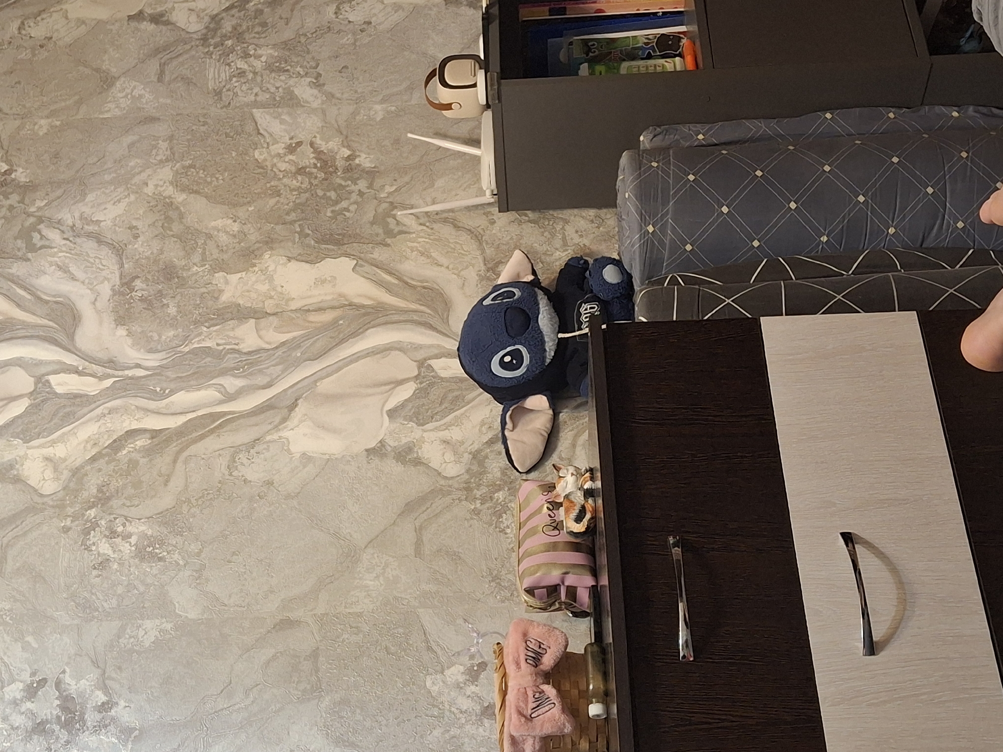 An indoor, eye-level, full shot shows a dresser and a shelf unit against a textured, marbled wallpaper. On the dresser are a woven basket with pink fuzzy ear warmers, a striped makeup bag, and a small figurine of two cats. A plush Stitch doll is seated on the edge of the dresser. To the right of the Stitch doll, on the shelf unit, are two white routers, a small white and brown portable speaker, and a shelf filled with books and colorful folders. A dark grey rolled blanket with a diamond pattern lies against the shelf unit. The bottom portion of the image shows the front of a two-toned dresser with chrome drawer pulls. A human foot is visible in the lower right corner of the frame.