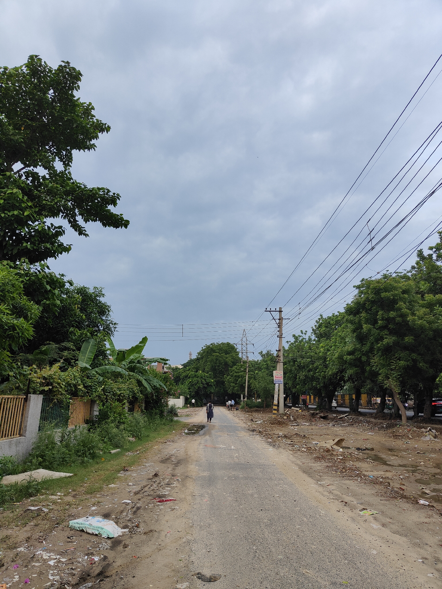 The image depicts a cloudy day scene on a rural road in Gurgaon, India. The road appears unpaved in some sections, with tire tracks and puddles of water indicating recent rain. The left side of the road is lined with dense vegetation, including trees and what appears to be banana plants, bordered by a yellow metal fence and a concrete wall. Scattered debris and a large white bag are visible on the ground in the foreground.
The right side of the road features a row of lush green trees, a utility pole with power lines stretching across the sky, and more debris along the roadside. A sign on the utility pole reads "TO-LET". Further down the road, construction or renovation activity is suggested by stacks of materials. Several people are walking along the road in the distance, and a person in dark clothing is walking away from the camera on the right side of the road. Cars are partially visible on the right side, indicating a populated area, despite the somewhat unkempt appearance. The overall impression is one of an urban fringe or a developing area within Gurgaon.