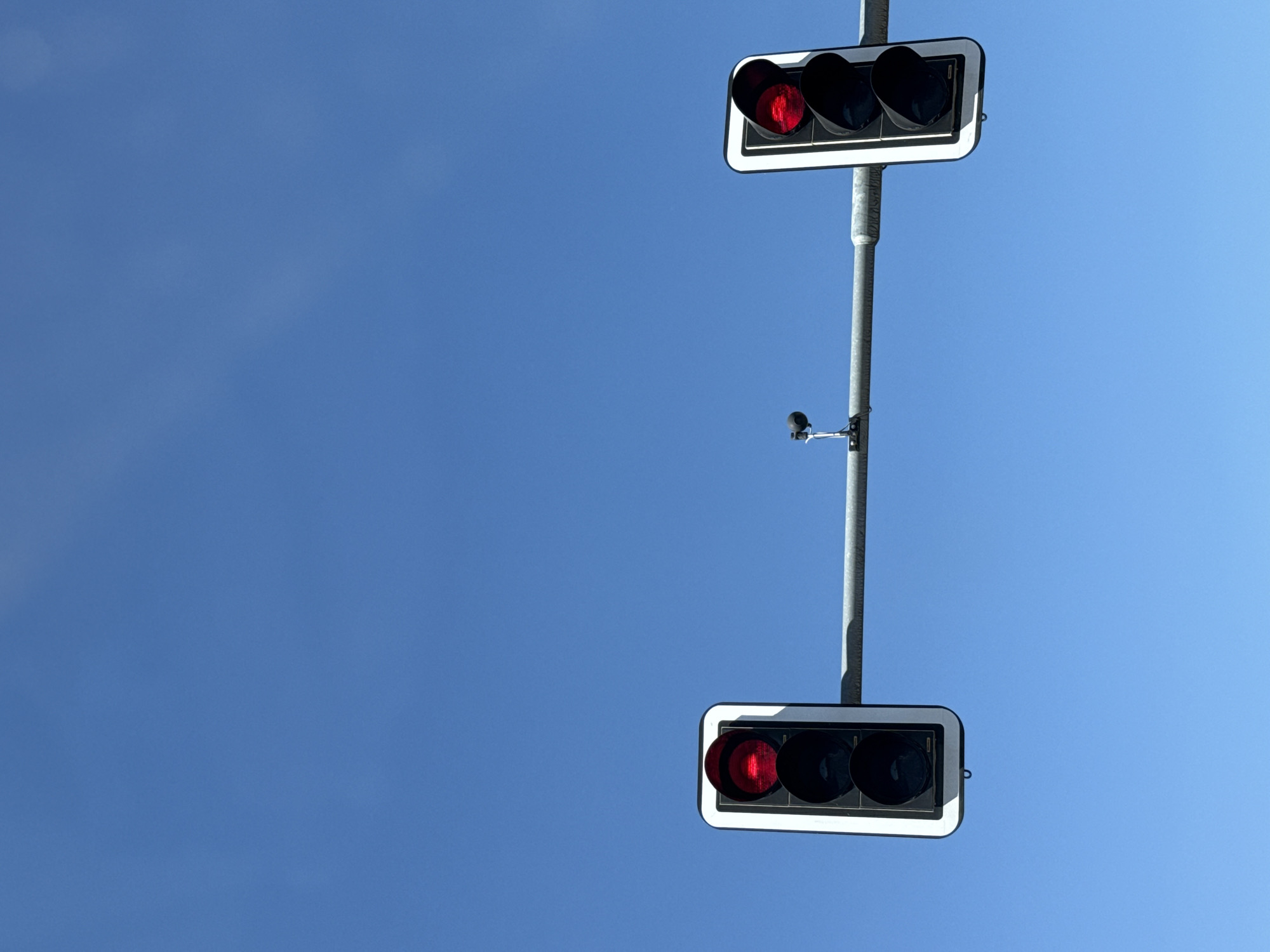The image shows two traffic lights against a clear blue sky. Both traffic lights are red. The traffic light on the left is positioned in the lower left corner of the frame, and the traffic light on the right is positioned in the lower right corner of the frame. A metal pole spans horizontally across the frame, supporting both traffic lights. A small black object, possibly a camera or sensor, is mounted on the pole between the two traffic lights. The sky is bright and appears cloudless, suggesting daylight. The scene does not show any people, vehicles, or buildings, indicating it might be an isolated traffic signal. The location is identified as Herzberg, Germany.