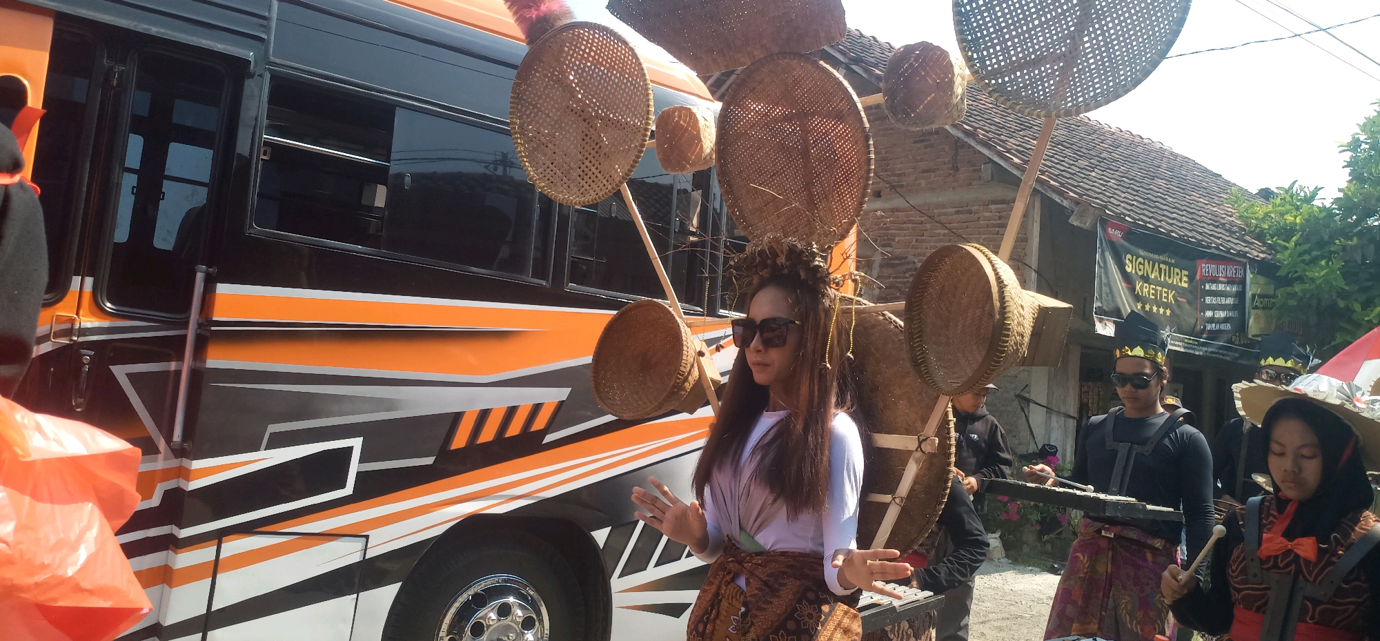 In the city of Kincang, Indonesia, a scene unfolds with a bus decorated in orange and black stripes. A woman with long brown hair, wearing sunglasses and a white top layered with a draped fabric and a patterned skirt, stands in the foreground. She has a headdress adorned with leaves and beads. Behind her, a collection of woven baskets of various sizes are suspended from poles, creating a unique installation.
To the right of the bus, several individuals are gathered. One person, wearing a black outfit and a golden crown-like headdress, plays a xylophone-like instrument. Another person, also in a black outfit with a similar headdress, stands beside them, holding drumsticks. Further to the right, a young person in a black outfit with a wide-brimmed straw hat and a red ribbon bow is also present, seemingly part of the musical group. Behind this group, a building with a tiled roof and brick walls is visible, and a banner with the words "SIGNATURE KRETEK" is displayed. The overall setting appears to be an outdoor event or festival.