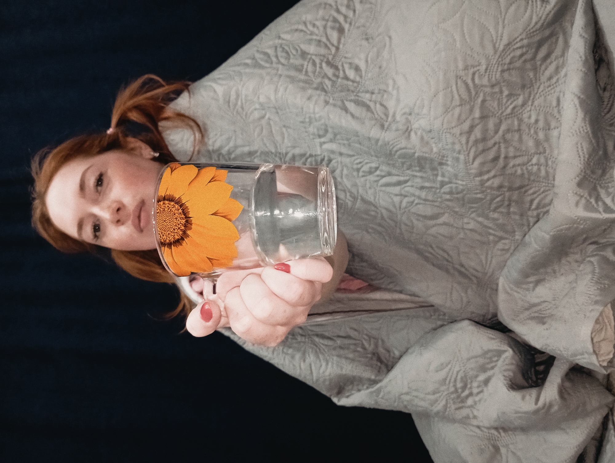 A woman with red hair is holding a clear glass with a sunflower sticker on it. She is wrapped in a gray patterned blanket against a dark blue background. The location is not discernible from the image.