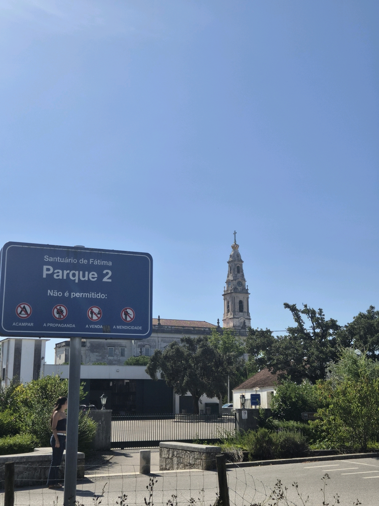 The image shows a blue sign with white text reading "Santuário de Fátima Parque 2. Não é permitido:" followed by four circular prohibition signs. The prohibition signs depict "ACAMPAR" (camping), "A PROPAGANDA" (propaganda), "A VENDA" (selling), and "A MENDICIDADE" (begging). In the background, a tall church tower with a cross on top is visible, characteristic of religious architecture in Fátima, Portugal. Green trees and buildings surround the area. A person is standing on the left side of the frame, facing away from the camera. The scene appears to be an outdoor area with a road in the foreground and a fence. The sky is clear and blue.
