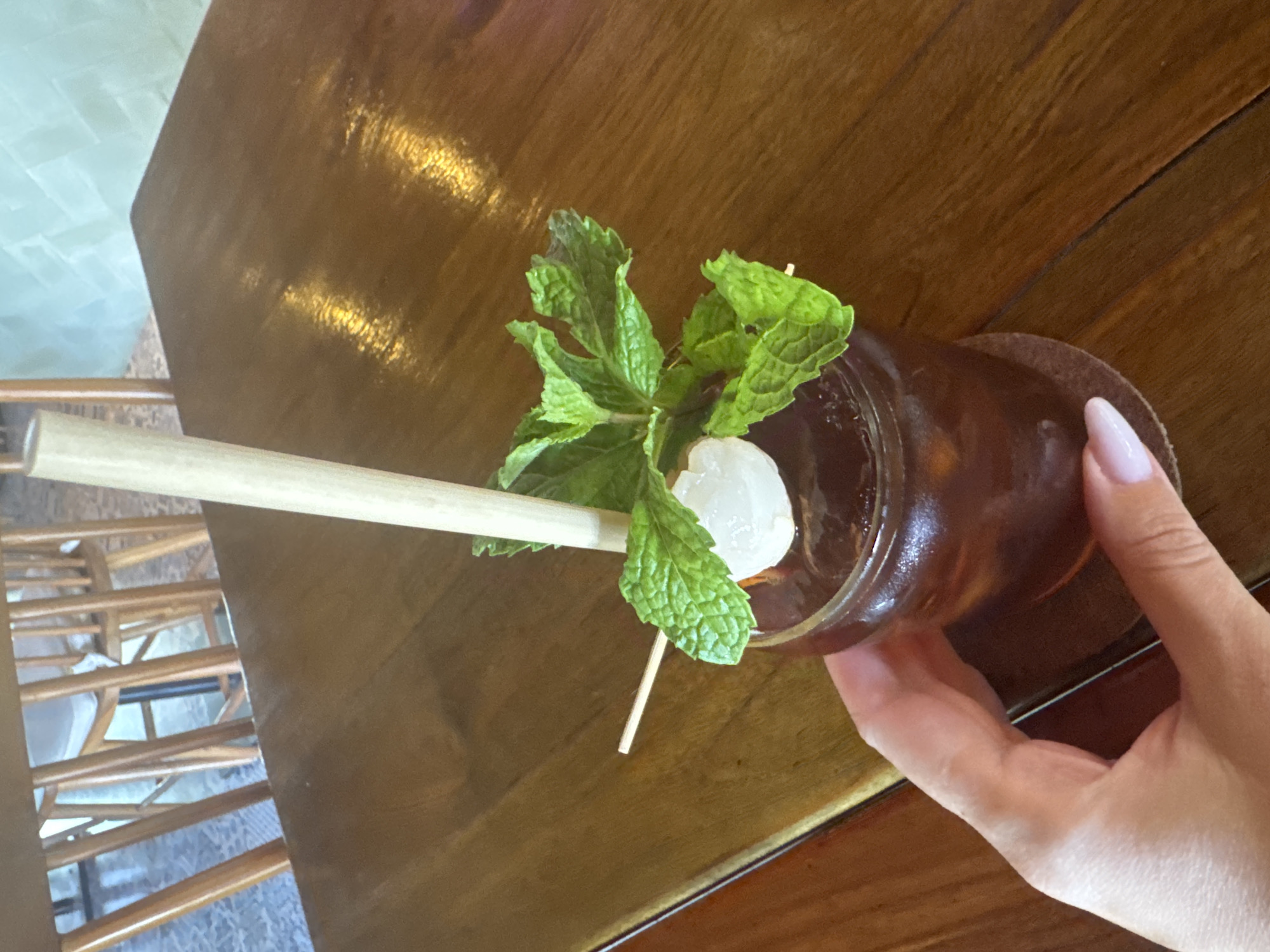 The image captures a close-up, high-angle shot of a beverage on a wooden surface. A brown-hued drink is visible, garnished with a sprig of mint and a lychee fruit, skewered by a wooden toothpick. A straw lies horizontally, extending from the beverage. A hand is holding the drink. The wooden surface is reflective. In the background, there is a partial view of wooden furniture, suggesting a cafe or restaurant setting. While it is not possible to determine the location in Denpasar, Indonesia definitively from the image, the presence of wooden furniture suggests a tropical or Indonesian-style cafe or restaurant.