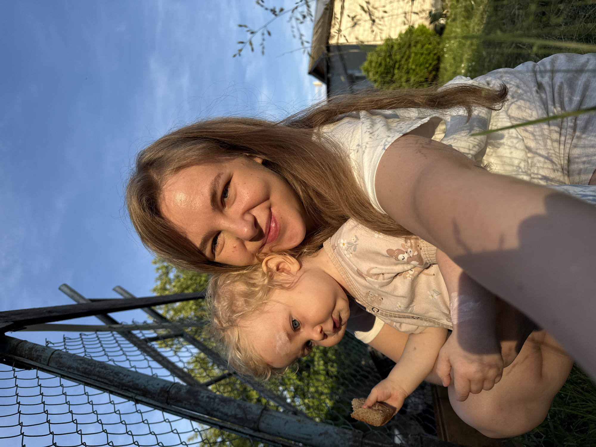 An outdoor portrait captured in a tilted selfie features a young adult woman embracing a baby. The woman, with long brown hair, smiles warmly at the camera. She wears a white top. The baby, with curly blonde hair and blue eyes, is held close to the woman, looking slightly off-camera. The baby has some food residue on its face and holds a small piece of bread in its hand.
The scene is set in what appears to be a garden or backyard in Красный Маяк, Russia. A metal chain-link fence runs across the mid-ground on the left side of the frame. Beyond the subjects, green foliage, including grass, bushes, and tree branches, is visible, along with parts of a building (possibly a house or shed) in the upper right background. The sky is clear and blue, with a faint contrail visible.
The lighting suggests late afternoon or early evening, often referred to as the golden hour, characterized by warm, soft sunlight. This light casts distinct shadows of leaves and branches onto the woman's arm and white dress. The overall mood is affectionate and joyful. No visible text is present in the image.