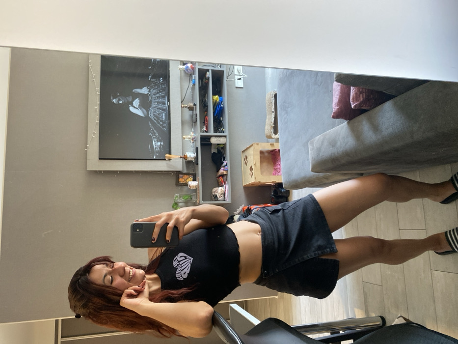 A woman with auburn hair smiles as she takes a selfie in a mirror. She wears a black crop top and a denim skirt. The reflection shows a gray-toned room with a television, decorative items on shelves, and a gray sofa. The flooring is light-colored tile, and she's sitting on a black chair. The room has a modern aesthetic with muted colors.