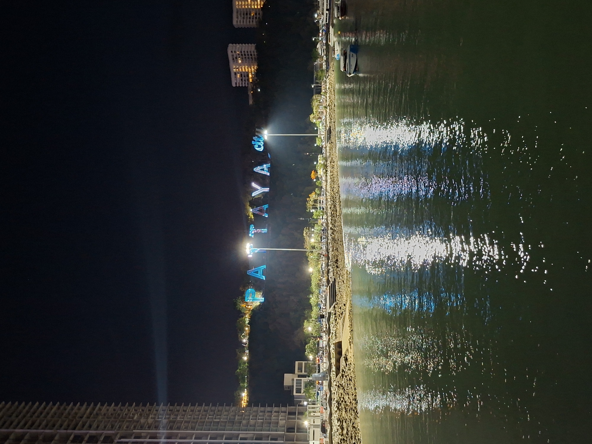 At night, a waterfront area in Pattaya City, Thailand, features the illuminated "PATTAYA" sign. A mix of dark sky and illuminated buildings is visible. The water reflects the city lights and the signs, creating a shimmering effect. Along the shoreline, there are buildings and streetlights. The foreground shows the edge of a building under construction.