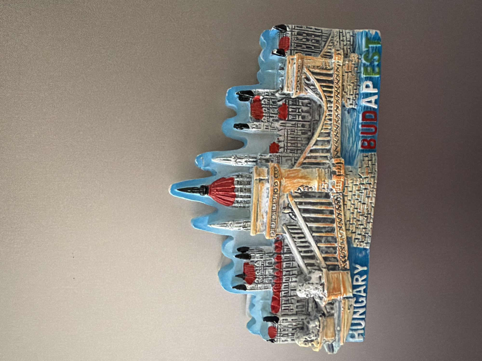 This is a close-up, eye-level, full shot of a refrigerator magnet depicting a miniature cityscape with the words "HUNGARY" and "BUDAPEST" on it. The magnet is horizontally oriented and shows a detailed representation of buildings, a bridge, and water. The architecture features red roofs, white walls, and some ornate details. The sky above the cityscape is depicted in a wavy, light blue pattern. The magnet is affixed to a plain, beige surface. There are no people or animals visible in the image. The background is a textured, neutral surface. The image is well-lit, with the magnet being the primary subject. There is no indication of Pazardzhik, Bulgaria in the image.