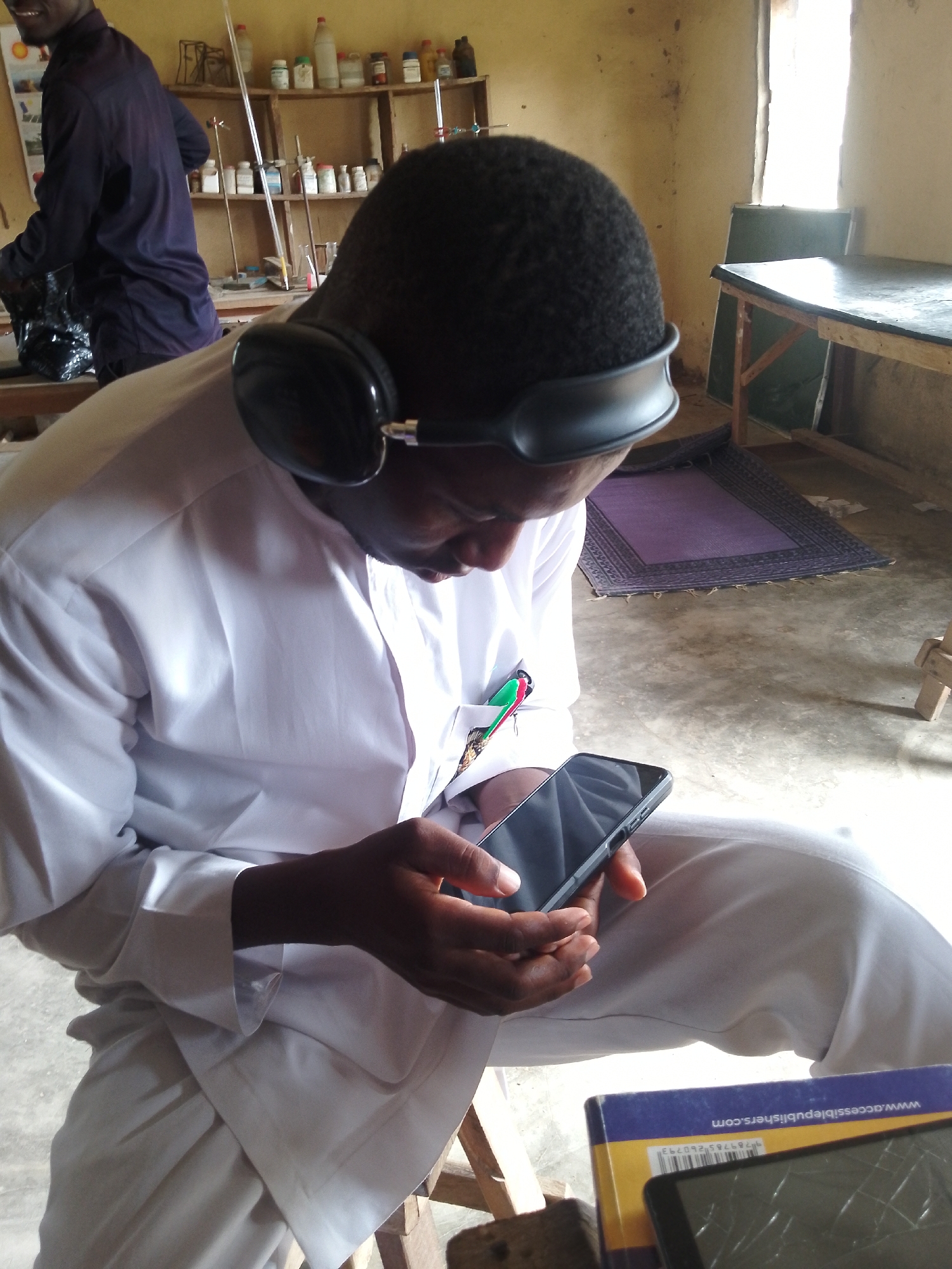 In a room that appears to be a laboratory or classroom in Serti, Nigeria, a young Black male wearing a white tunic and pants sits with headphones on, looking at a black smartphone. The room has shelves stocked with bottles and scientific equipment, and a wooden table with a black surface is visible near a window. Another person, wearing a dark purple shirt, is partially visible on the left side of the image, holding a black plastic bag. A purple rug is on the floor.