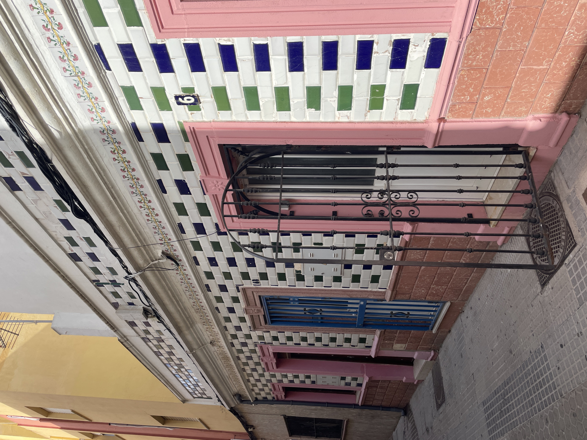 The image shows a narrow street in La Línea de la Concepción, Spain. The street is paved with gray cobblestones. On the right side of the street is a building with a facade covered in white, green, and blue tiles. The building has pink trim around the windows and doors. There is a black metal gate in front of one of the doors. The building number "6" is visible on the facade. On the left side of the street is another building with a yellow facade. The sky is visible at the top of the image. The scene is brightly lit by sunlight, casting shadows on the street and buildings. There are no people visible in the image.