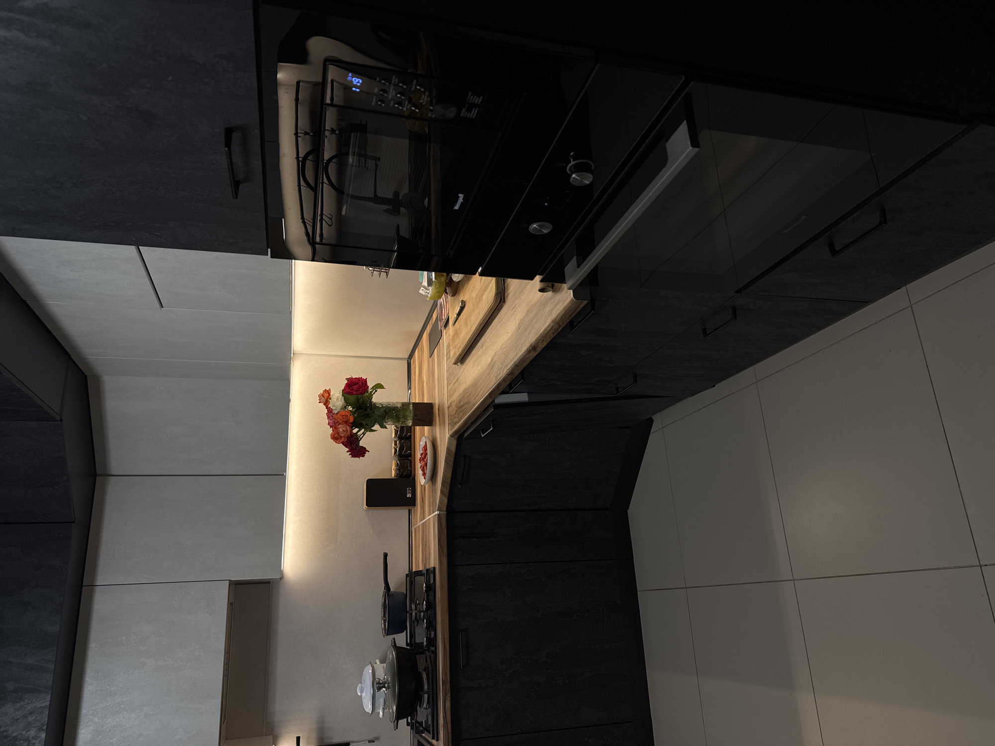 An interior view of a modern kitchen in Mikhaylovsk, Russia, shows a contemporary design with a mix of light and dark cabinetry. The countertop is a warm wood tone, contrasting with the dark, textured cabinets below. On the countertop, a gas stovetop features a large black pot with a lid and a smaller blue pot. To the right, a vase of colorful flowers, a small white plate with red berries, and a black speaker are arranged. A black built-in oven with a digital display is prominently featured on the right side of the frame. The floor is tiled with large, light-colored squares. Overhead lighting casts a warm glow on the backsplash and upper cabinets.