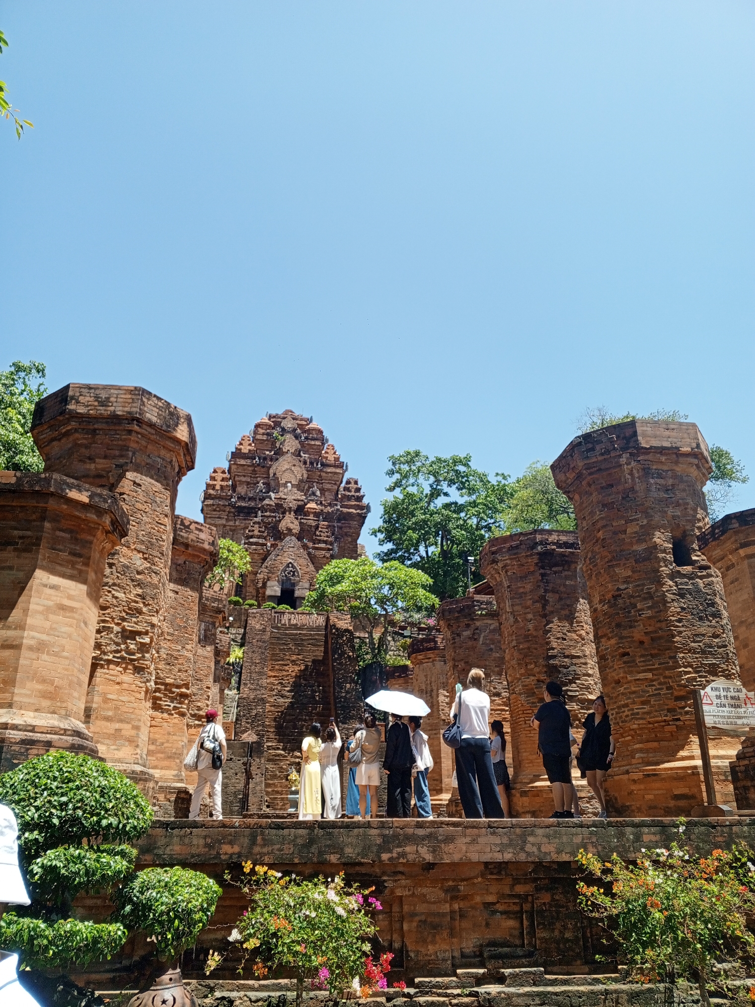 The image depicts the Po Nagar Cham Towers, located in Bac Nha Trang Ward, Vietnam, under a clear, bright blue sky. The scene is dominated by ancient, reddish-brown brick structures characteristic of Cham architecture.
In the background, a large, multi-tiered main tower stands prominently, featuring intricate carvings and architectural details, with a visible sign displaying "THAP BA PONAGAR" on its lower section. Surrounding this central tower are several tall, weathered brick pillars and walls, some appearing cylindrical or octagonal in cross-section. These structures exhibit signs of age, with textures of individual bricks and some minor vegetation growth.
A raised stone-paved platform or walkway extends across the mid-ground, where approximately ten to fifteen individuals, primarily adults, are present. Most people are dressed in casual attire, consistent with tourists. Notable individuals include: one person on the far left, partially visible, wearing a white hat; a person near the center holding a white sun umbrella; another individual in a white top and dark pants; a person wearing a white outfit and a red cap with a backpack; and two women standing closer to the main tower, one in a long yellow dress and another in a long off-white dress. Several individuals are engaged in observation or photography using smartphones. On the far right, a yellow warning sign is visible, containing text in Vietnamese and English, indicating "KHU VUC CAO DE TE NGAI / HIGH PLACES ARE EASY TO FALL".
The foreground features lush green foliage and various flowering plants, including potted, manicured shrubs resembling bonsai and bushes with colorful blooms in shades of pink, red, orange, and yellow. The overall environment suggests an archaeological or historical site open to the public during daylight hours.