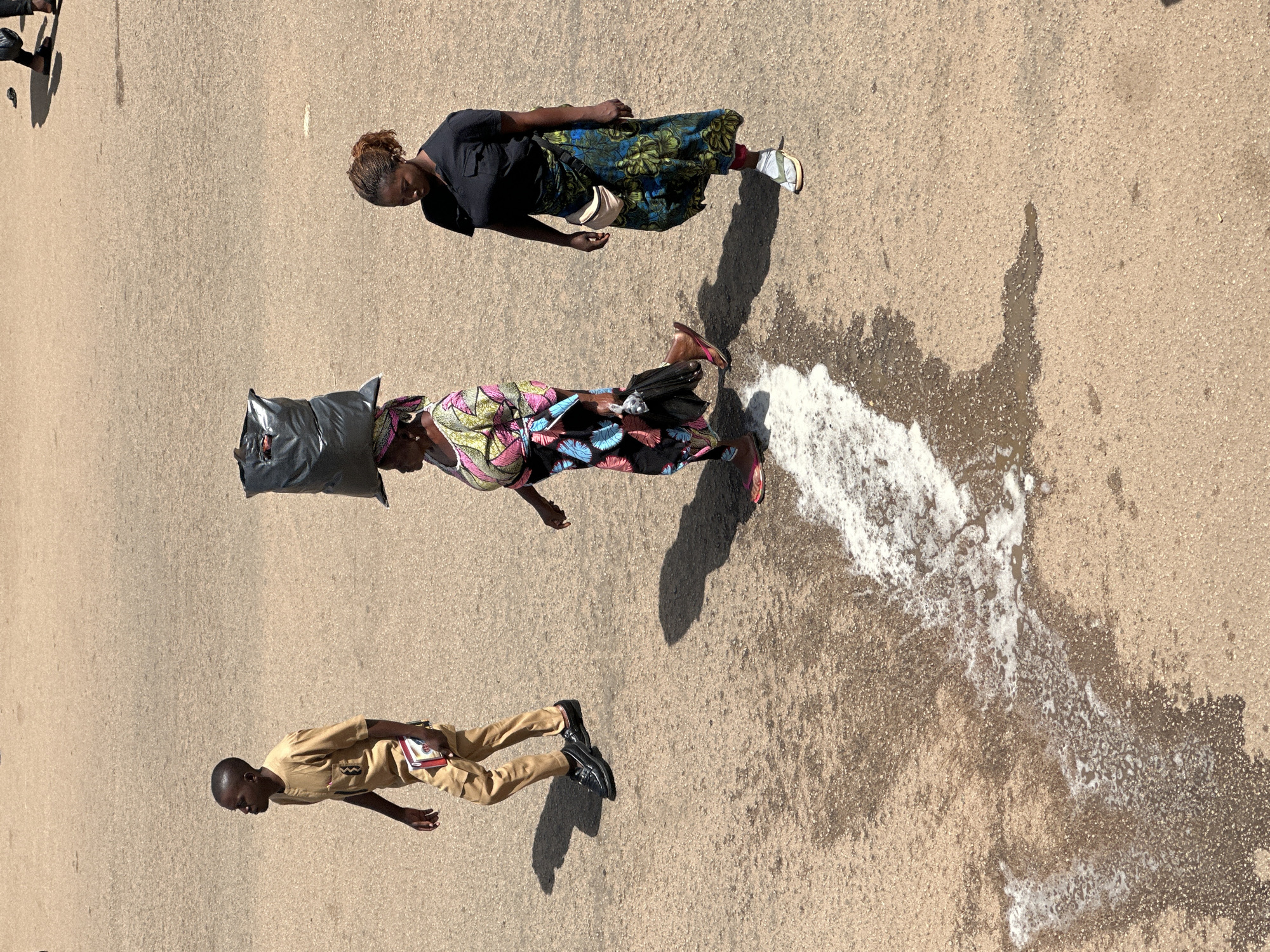 In Bukuru, Nigeria, three individuals walk across a dusty, paved surface under harsh sunlight. To the left, a young man in a tan outfit walks while holding a small book or notepad. In the center, a woman with a headscarf balanced a large black trash bag on her head. She wears a colorful, patterned dress and carries a black umbrella in her right hand. To the right, a woman in a black shirt and a long, patterned skirt walks, with a small beige fanny pack around her waist. A patch of white, foamy liquid, possibly detergent or soap suds mixed with water, is visible on the ground in the foreground, creating a wet, textured area. Shadows are cast sharply on the ground, indicating a bright, sunny day.