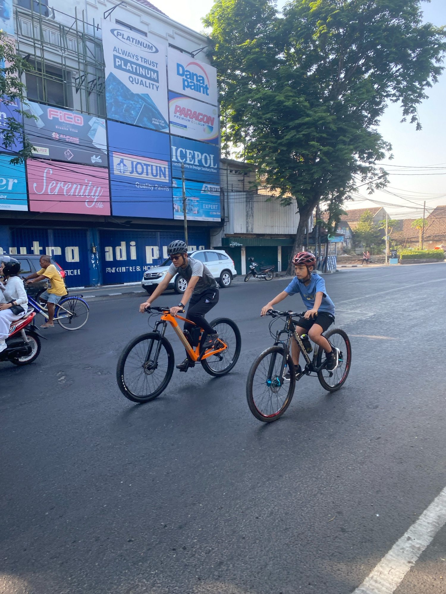 Two males are cycling on a street in Kediri City, Indonesia. The person in the foreground is wearing a helmet, sunglasses, a grey t-shirt, and black pants, and is riding an orange bicycle. The person behind him is wearing a helmet and a blue t-shirt, and is riding a black bicycle. They are both cycling in the same direction on a paved road. Buildings with advertisements are visible on the left side of the road. A tree with green foliage is on the right side of the road. Other vehicles, including a motorcycle and cars, are also present on the road. The sky is clear and blue.