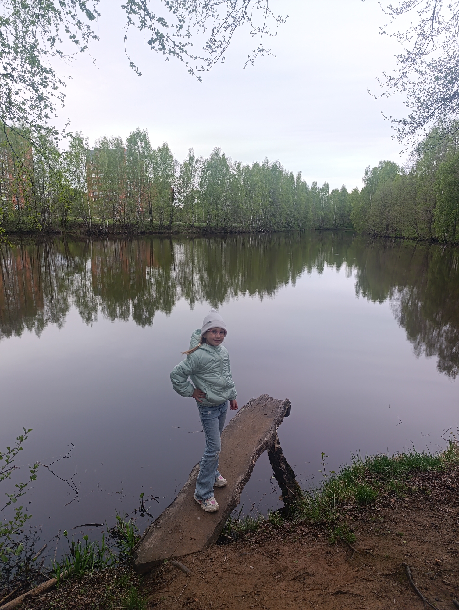 The image shows a young girl standing on a weathered wooden plank extending into a body of water. The girl is wearing a light blue jacket, jeans, and a white hat. She is looking at the camera with her hand on her hip. The body of water, likely a pond or lake, reflects the surrounding trees and a building in the background. The environment is natural, with trees lining the water's edge and some foliage in the foreground. Based on visual cues and the prompt specifying Chernogolovka, Russia, the image was likely taken in a park or recreational area within that city.