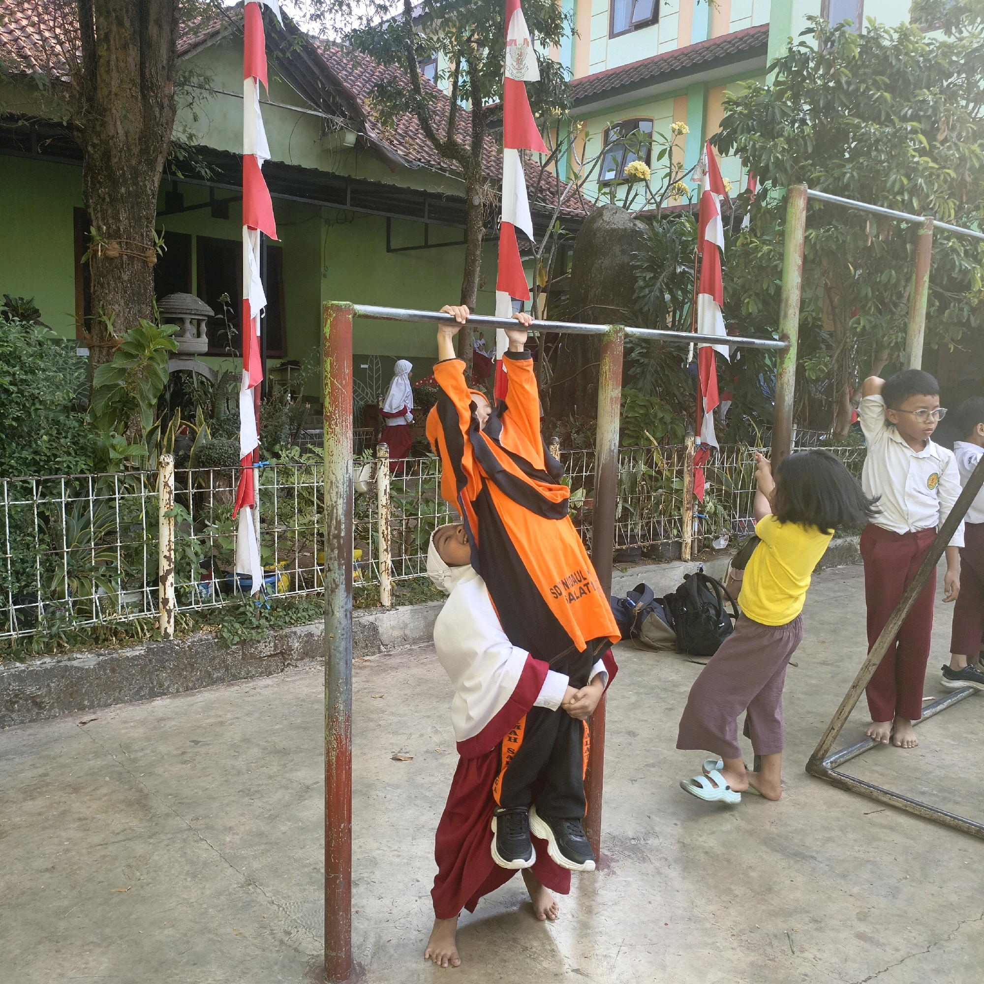 The image depicts an outdoor scene in Salatiga, Indonesia, likely at a school due to the presence of children in uniform and a building that appears to be a school structure. The foreground shows several children engaged in physical activity around a pull-up bar. Two children are actively interacting with the pull-up bar; one, wearing an orange and black uniform with a hijab, is hanging from the bar while being lifted by another child in a white and maroon uniform. Another child in a yellow shirt and brown pants is to the right, looking towards the children on the bar. A boy in a white shirt and maroon pants, wearing glasses, stands to the far right, his arm raised. The ground is paved concrete, and a metal fence with overgrown vegetation is visible behind the children. Red and white Indonesian flags are strung up, suggesting a national celebration or event. The building in the background is a light green structure with red tile roofing and windows. The overall atmosphere appears to be one of playful activity and camaraderie among the children in a schoolyard setting.