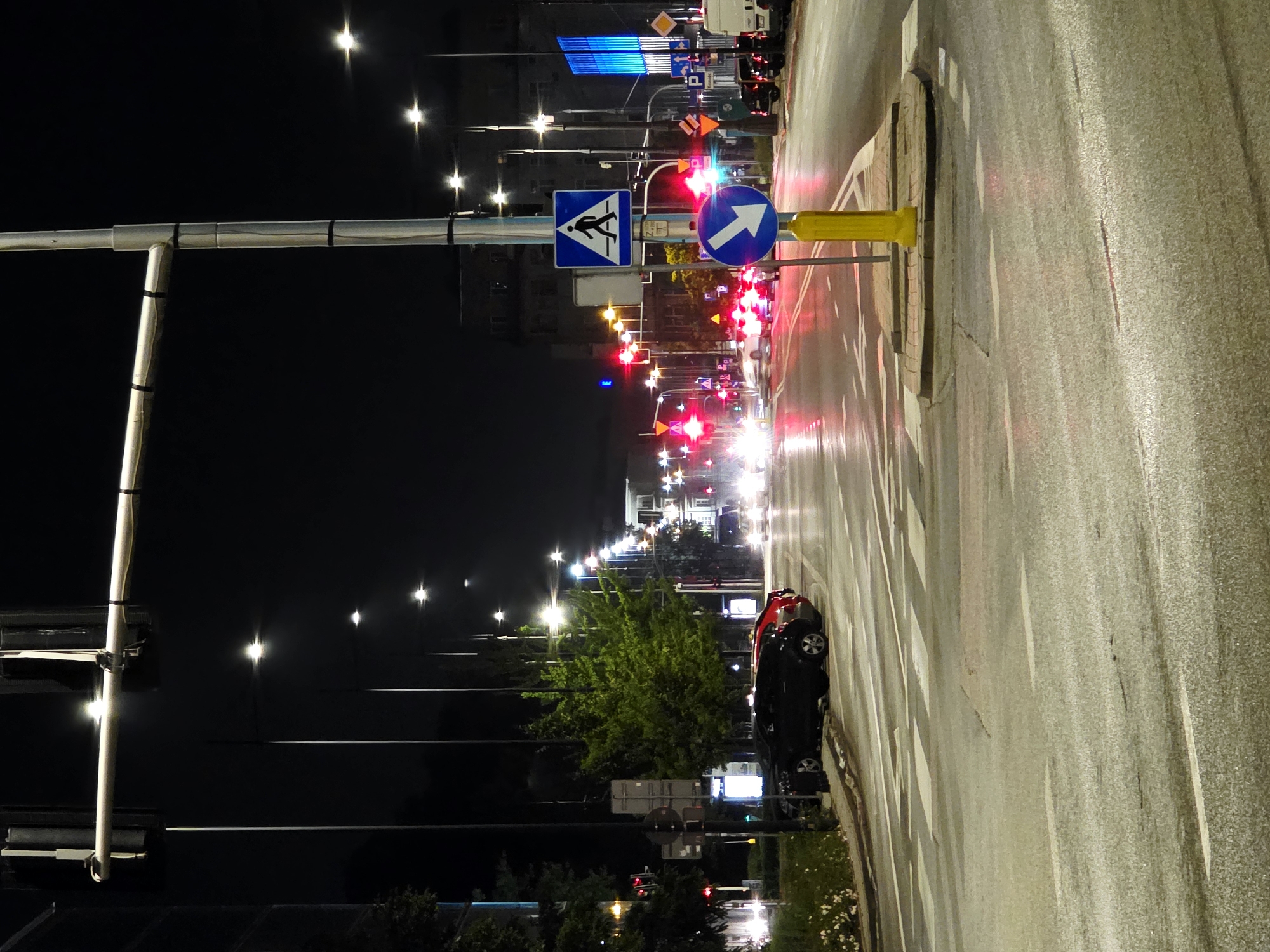 The image depicts a city intersection at night in Warsaw, Poland, likely in a commercial or mixed-use area. The scene is dominated by artificial lighting from streetlights, traffic signals, and illuminated buildings. A prominent element is a traffic light gantry spanning across the intersection, with multiple sets of traffic signals. Several cars are visible, some parked on the side of the road and others in motion, their headlights and taillights creating streaks of light. Pedestrian crossings are clearly marked on the wet asphalt, suggesting recent rain or damp conditions. Road signs, including a pedestrian crossing sign and a directional arrow sign, are present. Trees and foliage line the roadside in some areas. Buildings with illuminated windows are visible in the background, indicating urban density. The overall atmosphere is that of a typical urban environment at night, with traffic and infrastructure in operation.