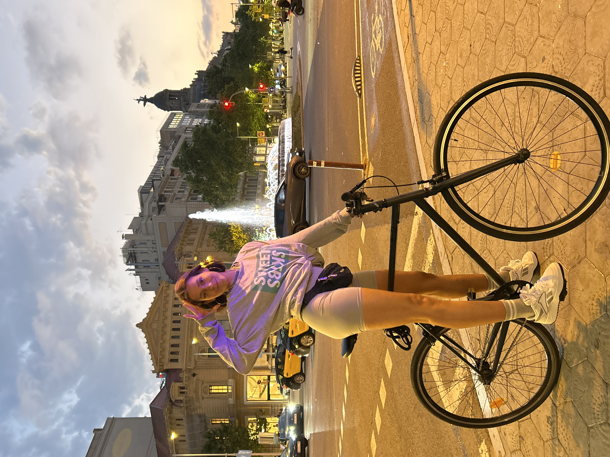 In Barcelona, Spain, a young woman is pictured with her bicycle at a street intersection during dusk. She is positioned to the left of the bicycle, with her left hand raised to her forehead in a "peace" sign gesture. She is wearing a light gray long-sleeved sweatshirt with green lettering that reads "SWEET SOXS," light gray bike shorts, white socks, and white athletic shoes. A black fanny pack is worn around her waist.
The bicycle is black and has a chain, pedals, and wheels with spokes. The woman's legs are visible, with one foot resting on a pedal. The background shows a fountain, several buildings with architectural details, streetlights, traffic lights, and a few vehicles, including cars, a taxi, a scooter, and a motorcycle. The sky is overcast with clouds, and the overall lighting suggests the twilight hour, with streetlights and building lights illuminating the scene. The ground is paved with interlocking hexagonal tiles.