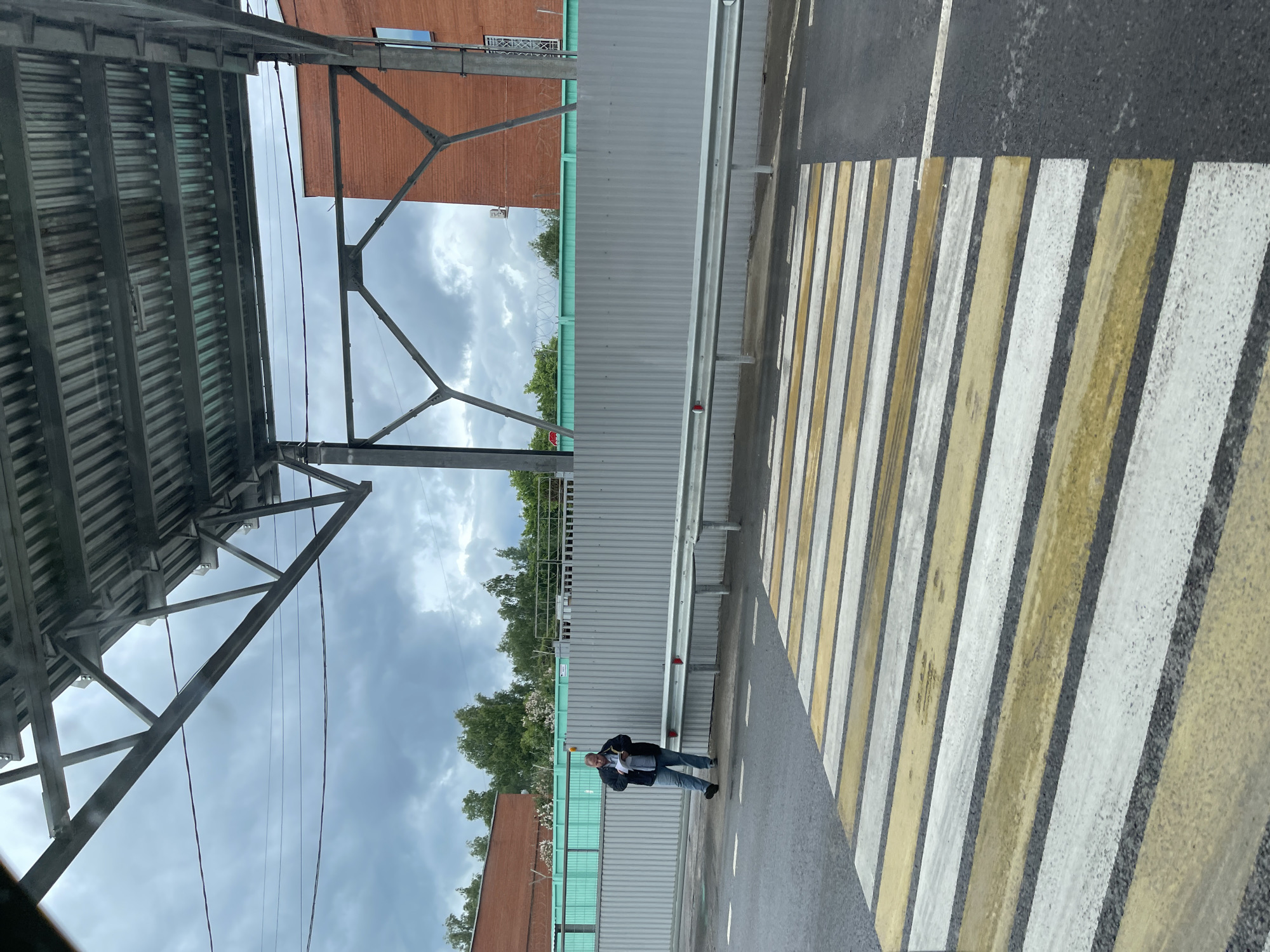 The scene is set in the city of Taraskovo, Russia. A person is standing on a crosswalk. Overhead is a metal structure, possibly part of a covered walkway or bridge. A gray corrugated fence is also visible in the background. Beyond the fence are trees and buildings. The sky is overcast.