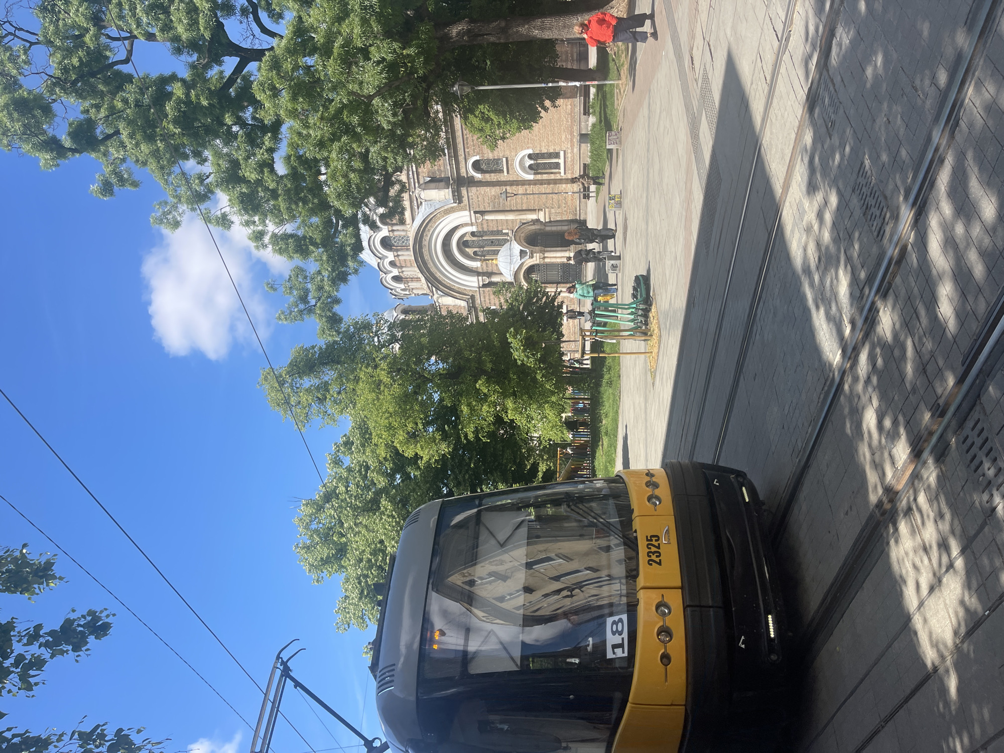 The image shows a street scene in Sofia, Bulgaria, featuring a yellow and black tram with the number 2325 and 18 prominently displayed. The tram is positioned on tracks running along the street. In the background, the Saint Sofia Church is visible, characterized by its arched windows and dome-like structures. A person wearing a red jacket is walking on the sidewalk near the church. Trees are present, adding greenery to the urban landscape. The sky is blue with some clouds, indicating a sunny day. People are standing near the church, some with scooters. The image captures a moment of everyday life in Sofia.