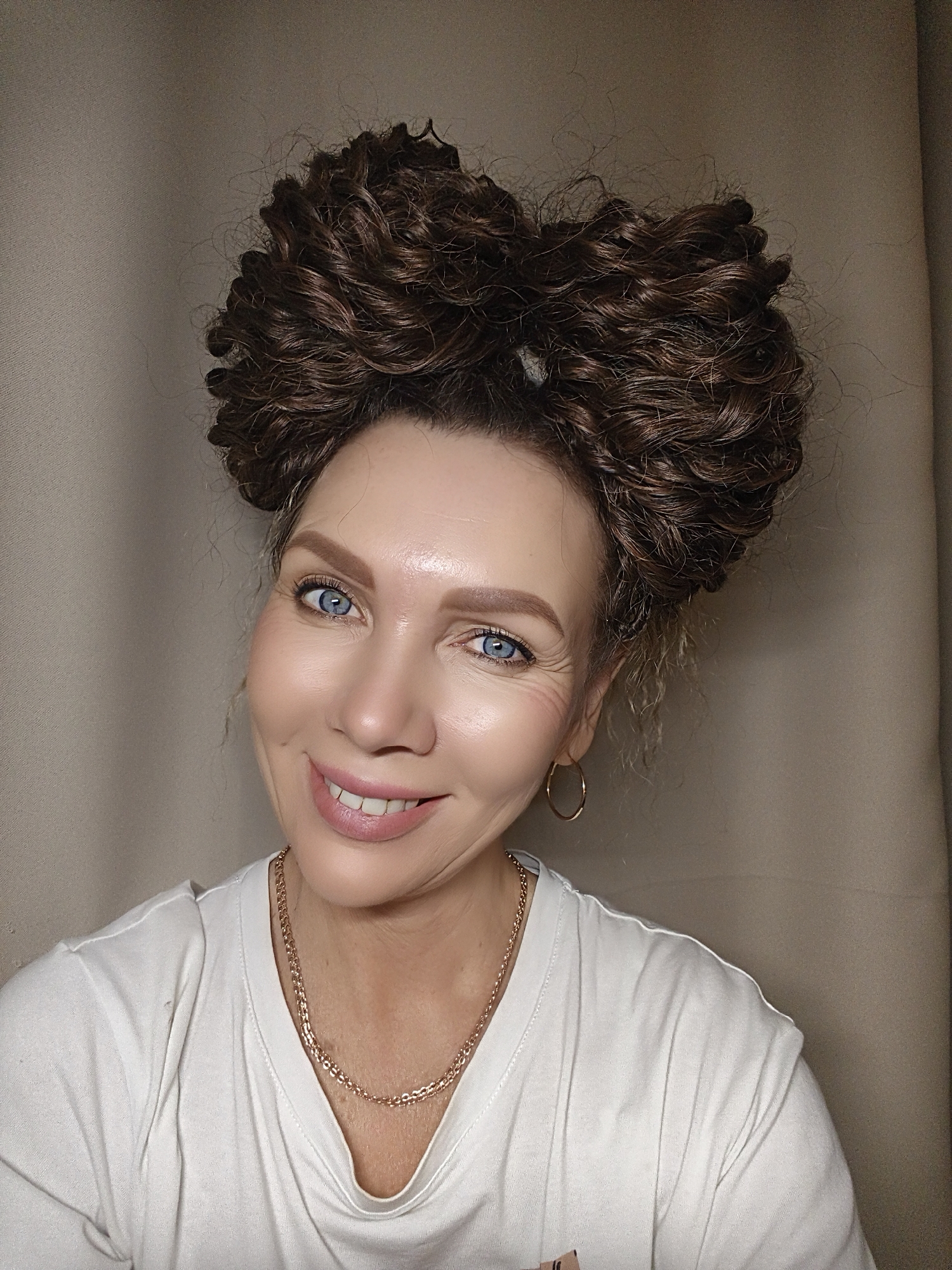 The image displays a close-up photograph of a Caucasian woman, appearing to be middle-aged, positioned from the chest up and centered. She exhibits a broad smile, revealing her upper teeth, with her blue eyes wide open and crinkling at the outer corners. Her eyebrows are well-defined and brown. Her dark brown, curly hair is styled into two large, distinct buns on top of her head, resembling a bow. She is wearing gold-colored hoop earrings in both ears and a gold-colored chain necklace. Her clothing consists of a white, short-sleeved t-shirt. The background is a plain, light brown or beige textile surface, indicating an indoor environment within the city of Unknown, Unknown. The lighting is soft and appears to be uniformly distributed across the subject's face.