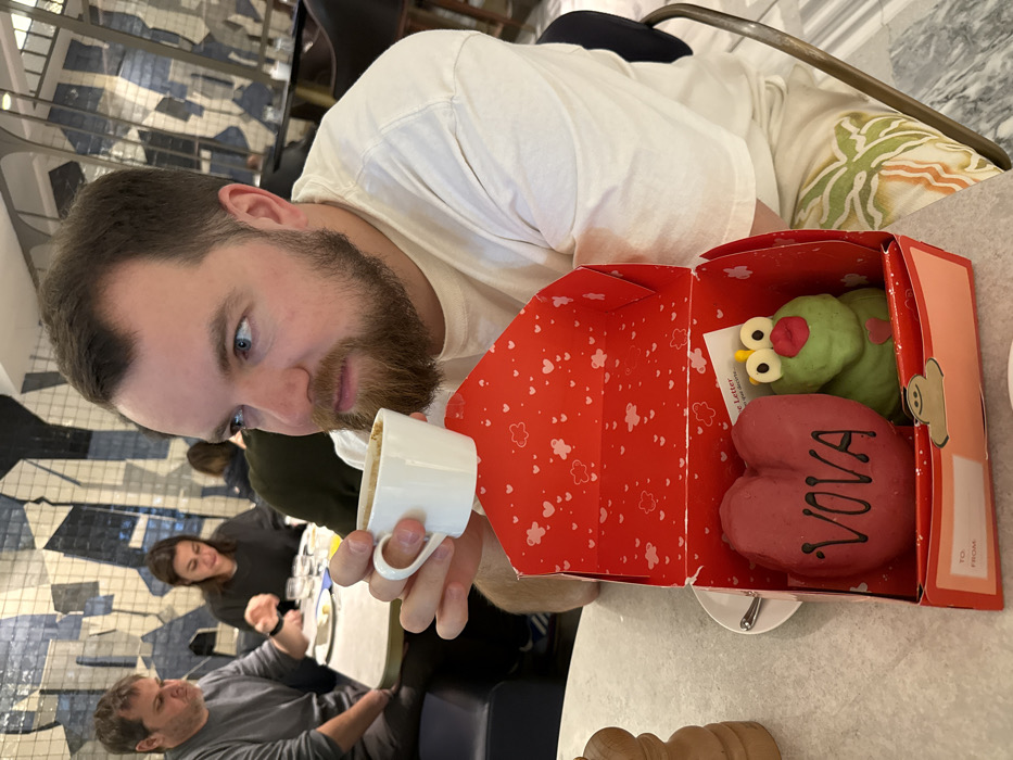 The image displays the interior of the Blanc restaurant located within the Mandarin Oriental Barcelona, Spain. In the foreground, a man with a beard and blue eyes, wearing a white t-shirt and light-colored shorts with a green leaf pattern, holds a small white espresso cup to his mouth. Positioned on the light-colored table directly in front of him is an open red gift box, patterned internally with white hearts and clouds, and featuring a "TO: ____ FROM: ____" label on its lower front flap. Inside the box are two custom pastries: a large red heart-shaped pastry with "VOVA" inscribed in white capital letters, and a green frog-shaped pastry with prominent white eyes, red lips, and a small yellow crown-like detail on its head, along with a small red heart on its chest. A wooden pepper grinder is partially visible on the left side of the table. In the midground, another man in a grey shirt and a woman in a dark top are seated at a separate table, engaged in dining. The background features a distinctive wall adorned with an abstract mosaic pattern in various shades of blue.