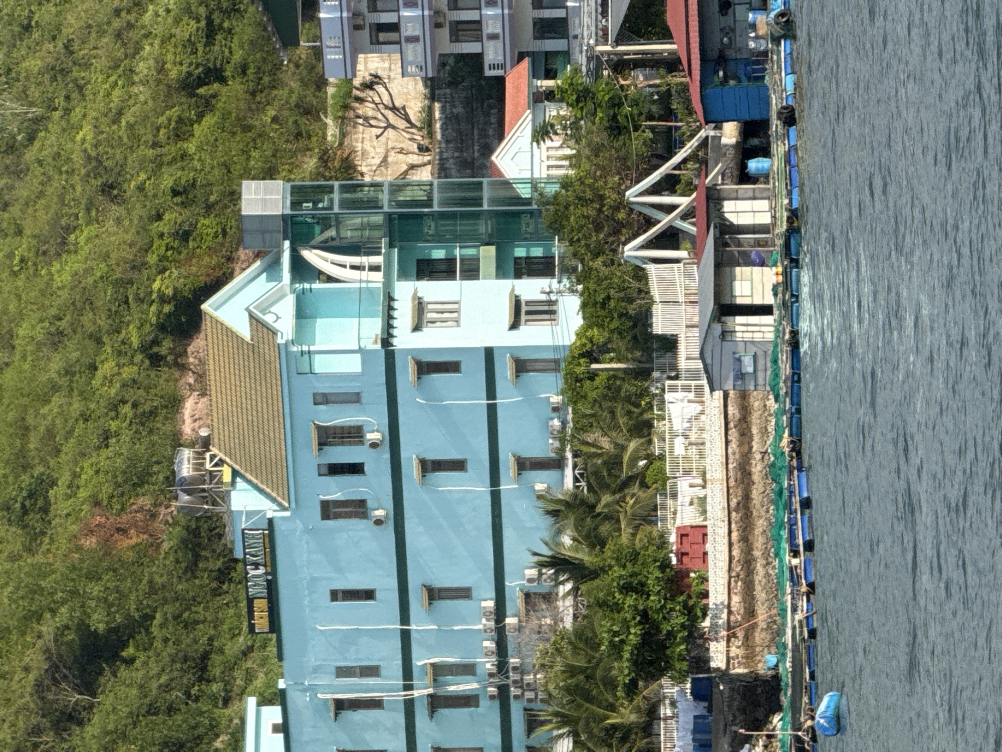 This is a photograph of a blue building situated on the coast, with a lush green hillside in the background. The building appears to be a hotel or guesthouse, with multiple floors and windows. There is a visible sign on the building that reads "NGOC XANH". In front of the building, there is a body of water, likely the sea, with a floating structure that could be a fish farm or dock. Several small structures, possibly utility buildings or sheds, are located on the floating platform. The overall scene suggests a tropical or coastal environment. The location is Nha Trang Ward, Vietnam. There are no people visible in the image.