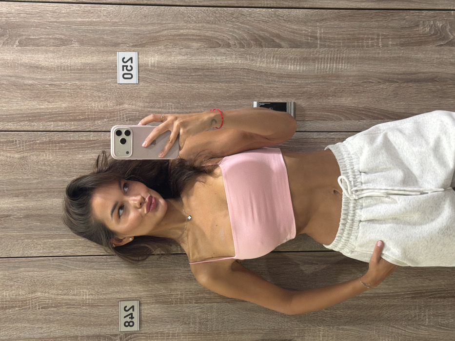 The image depicts a single female individual taking a mirror selfie within what appears to be a locker room environment. The background features light brown wooden locker doors, displaying reflective numerical labels "S48" and "S20" (as seen in reflection). A metallic lock mechanism is partially visible on the right-hand locker door. No specific identifying features are present to pinpoint the exact location within the city of Krasnoyarsk, Russia.
The subject is a woman with long, dark brown hair, styled with volume, partially obscuring the left side of her face. She is wearing a light pink, thin-strapped crop top, revealing her midriff, and light grey drawstring sweatpants. Her left hand is partially inserted into the pocket of her sweatpants. She is accessorized with a silver necklace featuring a small, four-leaf clover-like pendant, small stud earrings, a thin silver chain bracelet on her left wrist, and a red string bracelet with a crescent moon charm on her right wrist. Her right hand holds a light-cased smartphone, consistent with an iPhone model, pointed towards the mirror. The woman is looking directly at the phone's camera with a "puckered lips" expression.