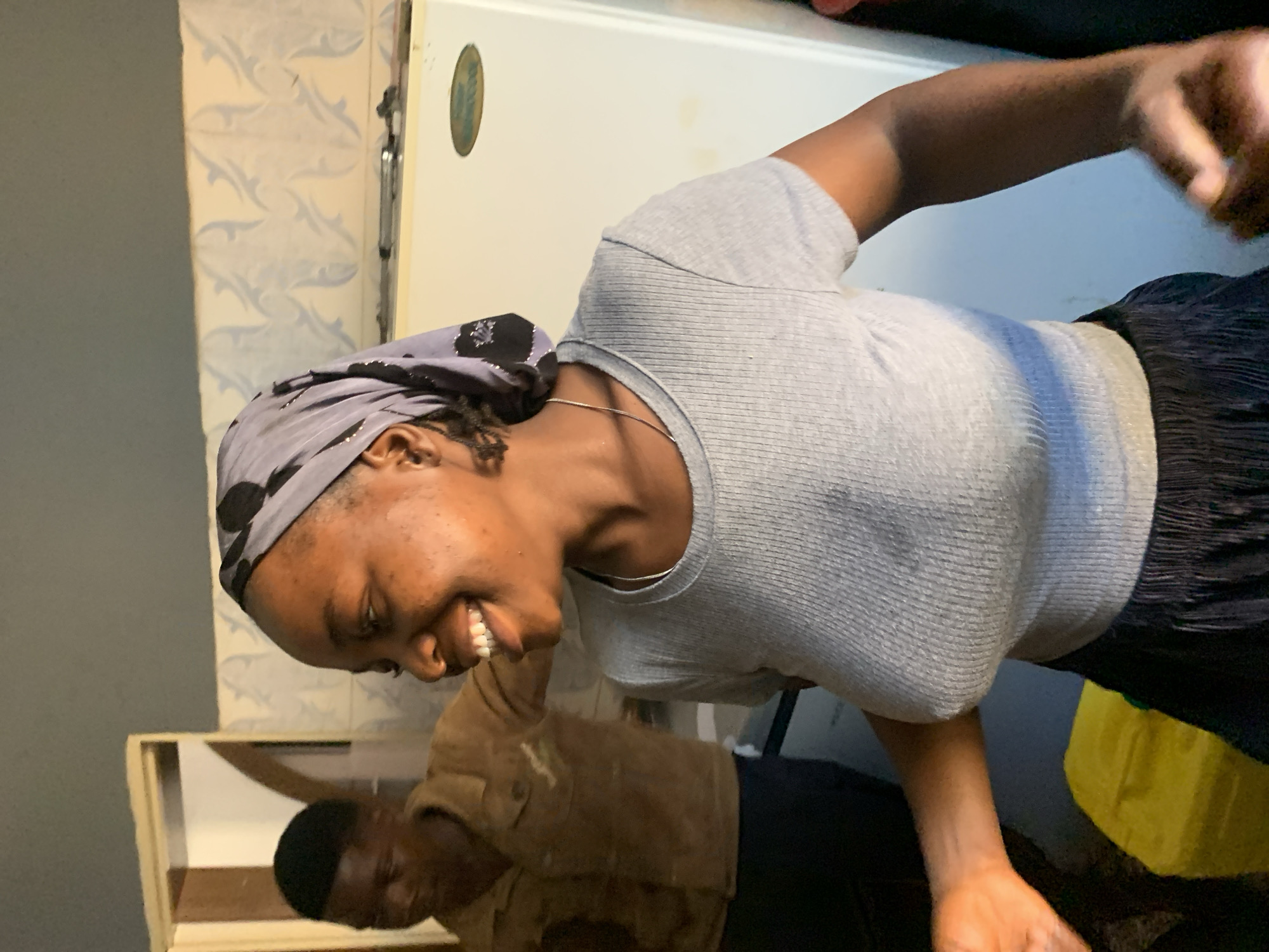 In an indoor setting, possibly a kitchen in Jos, Nigeria, a young Black woman is the primary subject, captured in a medium shot from the chest up. She wears a grey ribbed t-shirt and a grey headscarf with a black polka dot pattern. Her head is tilted to her left, and she has a broad smile, revealing her teeth. A thin silver necklace is visible around her neck. In the background, a man in a brown jacket stands to the left of the woman, partially obscured. The environment includes a light-colored tiled wall with a decorative pattern, and a white refrigerator to the right of the woman, which has a golden oval emblem on it. A doorway with a beige frame is visible on the far left. The lighting suggests an indoor environment, possibly with natural light contributing to the illumination.