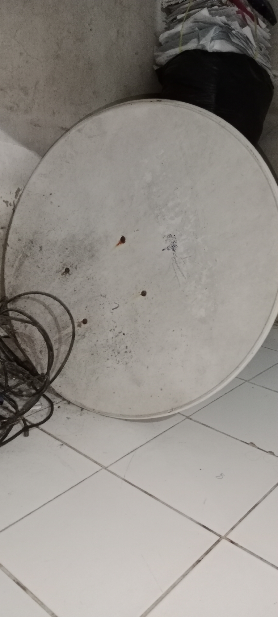 The image shows a satellite dish lying flat on a white tiled floor in a dimly lit interior, possibly a room or storage area in Subang, Indonesia. The satellite dish is round and off-white, with visible dust and some dark markings on its surface. Several screws are present on the dish. To the left of the dish, a coiled black cable lies on the floor. In the background, a wall with a rough texture is visible, and on the upper right, a black garbage bag filled with stacked papers can be seen. The overall scene suggests a utilitarian or storage space, possibly a garage or a backyard storage area. There are no people or animals present in the image.