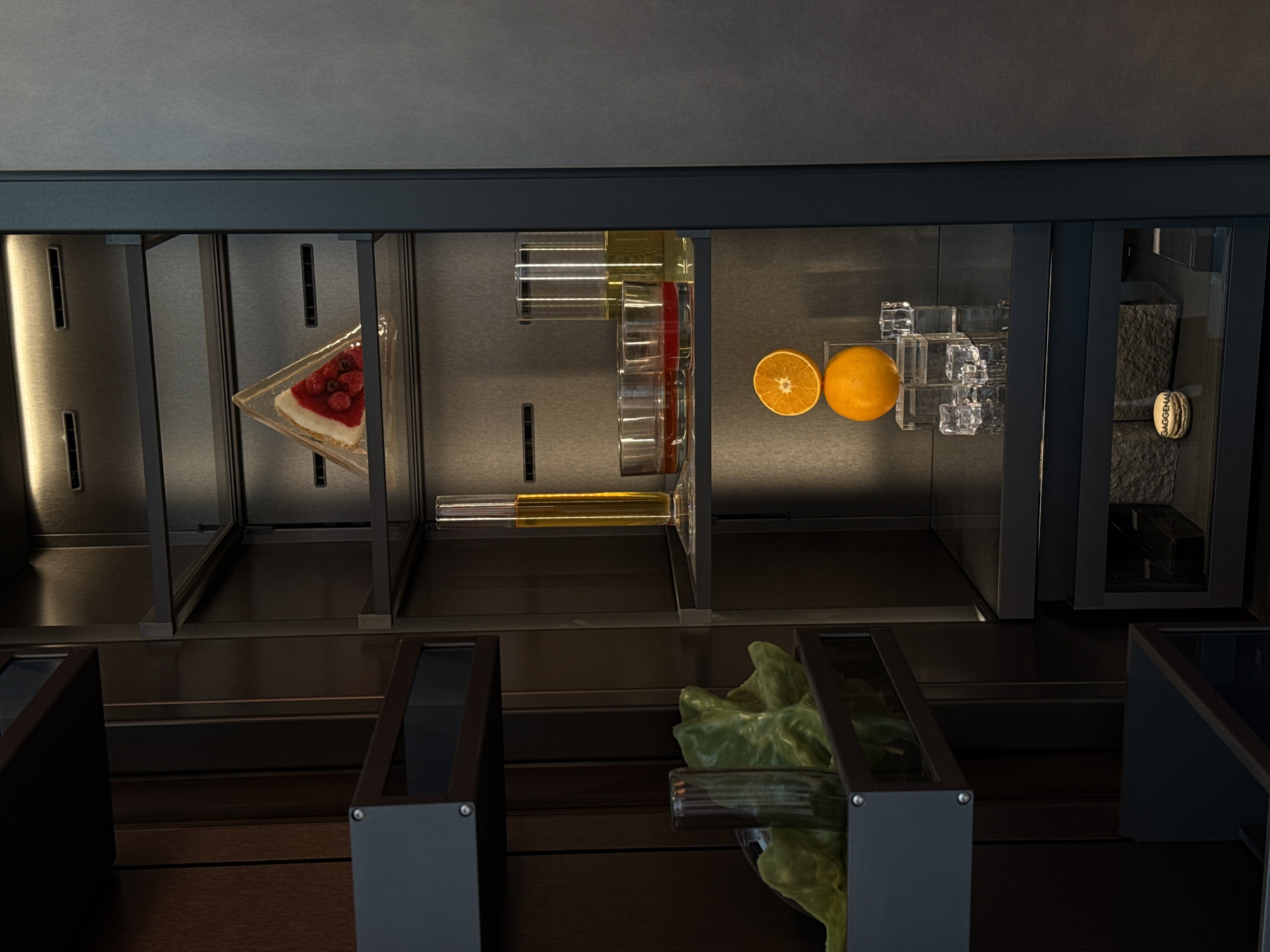 The image depicts a meticulously organized refrigerated display case, viewed from an elevated perspective. Inside, a variety of food items are arranged in separate compartments. From left to right, there is a triangular slice of cheesecake topped with red berries, several clear glasses containing liquids (possibly juice and water), an orange cut in half and a whole orange, several stacked glass cubes that resemble ice, and two stacked rough stones with a macaroon placed on top. In the front of the display case, slightly below the refrigerated section, there is a container holding green lettuce. The materials and the color palette indicate a sleek, modern design. The scene is likely set in a high-end grocery store or restaurant in Milan, Italy.