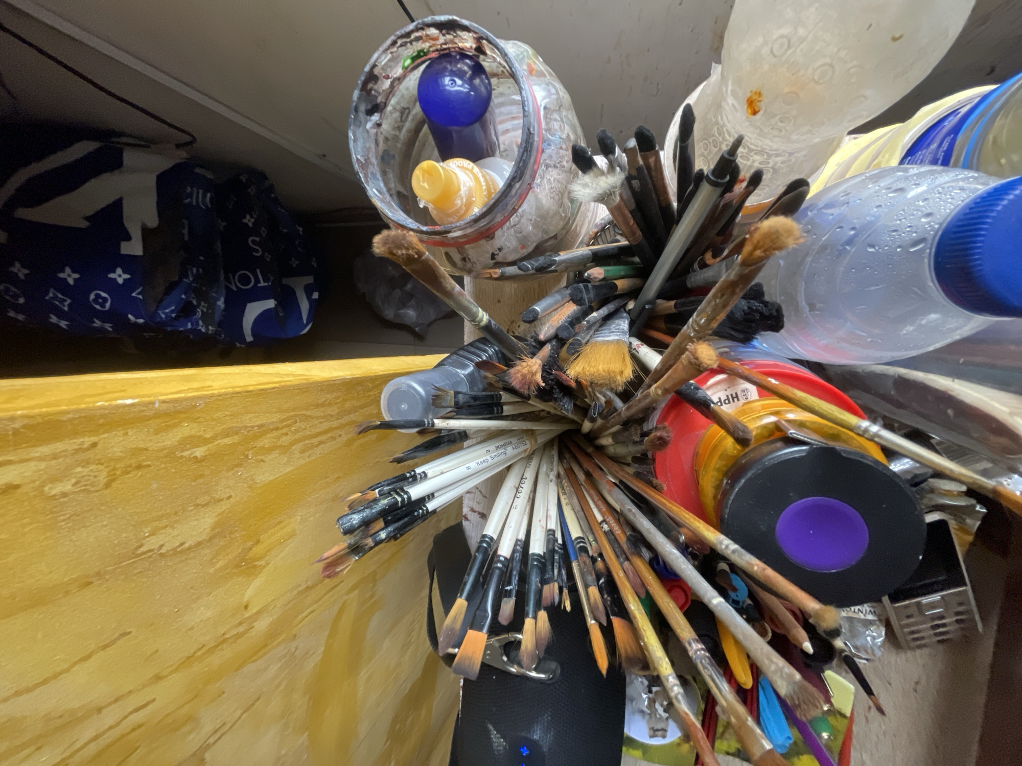 The image depicts a close-up, overhead view of an artist's workspace, likely in a home setting in Ado, Nigeria. A collection of paintbrushes is the central focus, clustered together in a container. The brushes vary in size and bristle type, with some appearing well-used and stained with various colors. Interspersed among the brushes are several containers and bottles, including a glass jar containing what appears to be water or cleaning fluid, a spray bottle with a yellow nozzle, and a round black container with a prominent purple lid. Several plastic bottles, one partially filled with water, are also visible. In the background, a blue fabric with a white pattern, possibly a throw or bag, is partially visible. The environment appears somewhat cluttered, suggesting an active and utilized creative space. The overall impression is one of a functional, perhaps somewhat informal, artist's studio.