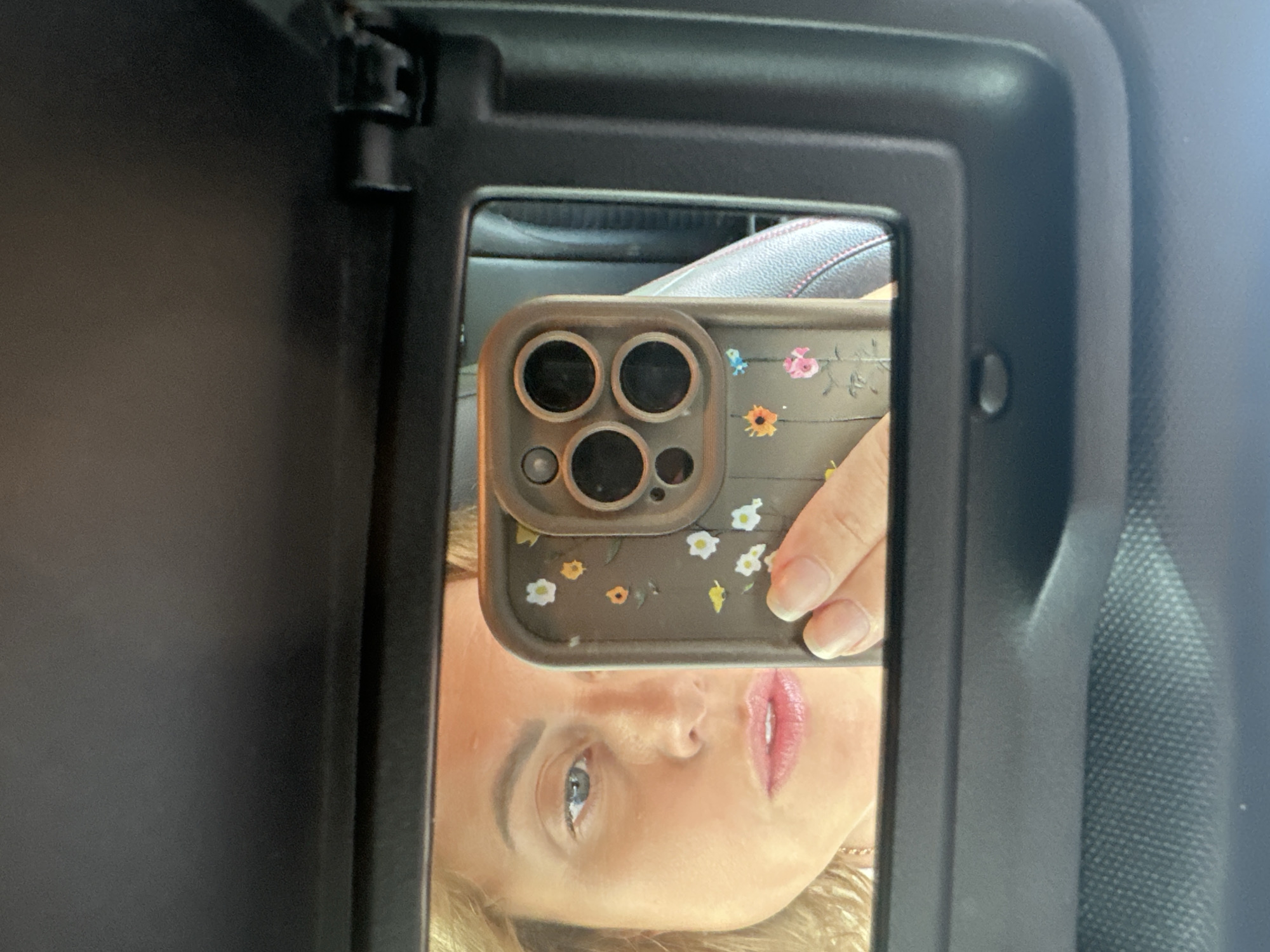 In a car's sun visor mirror, a woman's reflection is visible. She has blonde hair and is wearing makeup, with glossy pink lipstick and blue eyeshadow. Her left eye is looking directly at the camera, while her right eye is obscured by the phone she is holding up to take the picture. The phone has a floral patterned case with small daisies and sunflowers. The woman is wearing a necklace. The background is a blurred car interior, with a hint of a leather seat. The mirror is framed by a black sun visor, and a small button is visible below the mirror. The overall lighting suggests it is daytime, possibly sunny, with light reflecting on the woman's face and the mirror.