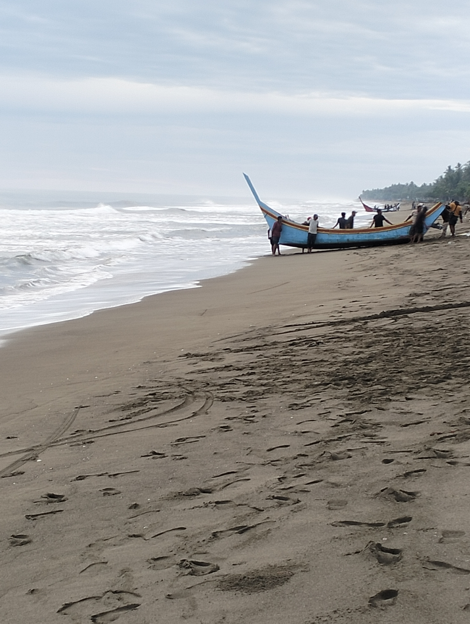 This image depicts a beach scene in Curehtunong, Indonesia, with a blue and yellow fishing boat being pulled ashore by several individuals. The boat, of traditional design, rests on the wet sand near the shoreline, where waves are breaking. The sky is overcast, and the background shows a line of palm trees. Numerous footprints are visible in the sand, indicating recent activity. The overall atmosphere is one of ongoing work related to fishing or boating.