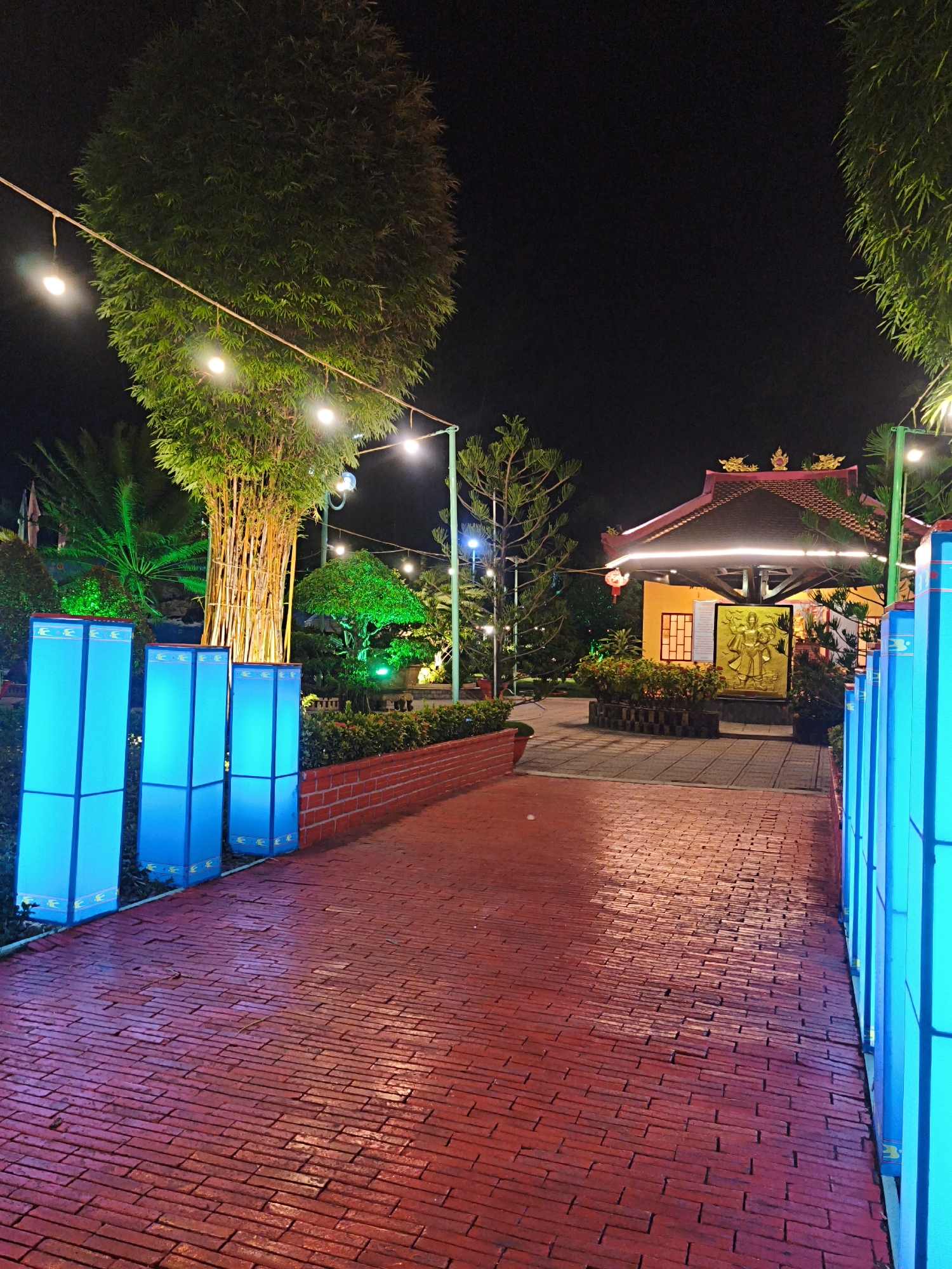 This is a nighttime photograph of an outdoor paved walkway lined with blue illuminated lanterns. The walkway is made of red brick and appears to be wet, reflecting the surrounding lights. To the left of the walkway, there are several tall, rectangular blue lanterns. Further back, a large bamboo tree stands illuminated by yellow light, and strings of fairy lights are visible above. To the right, the walkway continues and is also lined with blue lanterns. In the background, a building with a traditional East Asian architectural style, featuring a red tiled roof and yellow walls, is visible. A golden relief sculpture of figures is mounted on the building's facade. The environment is landscaped with trees and bushes, some of which are illuminated with green lights. The overall scene suggests a festive or decorative outdoor space, possibly a temple or cultural attraction, located in Bac Nha Trang Ward, Vietnam. No people are visible in the image.