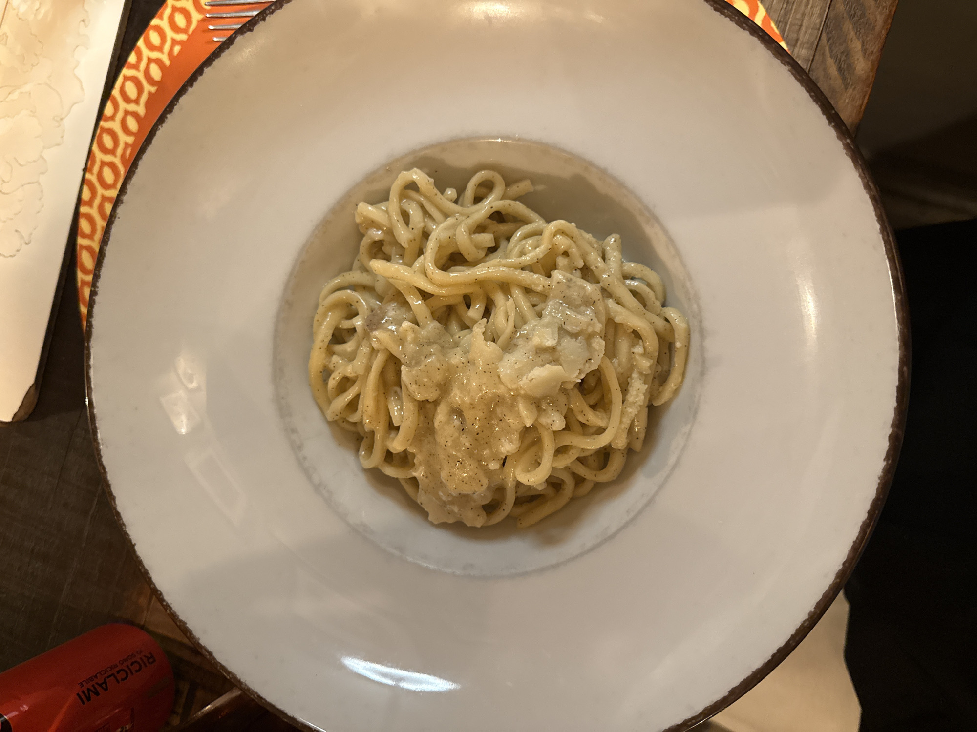 The image shows a plate of pasta, likely spaghetti or a similar type, served with a light-colored sauce. The pasta is arranged in a central well of a wide, white plate with a subtly textured rim. A partial view of a patterned orange and white surface is visible to the upper left of the plate. The edge of a wooden surface is visible beneath the plate, and part of a red can with Italian writing on it is also partially visible. The location is likely a restaurant in Rome, Italy, based on the presence of Italian writing on the can and the style of pasta dish.
