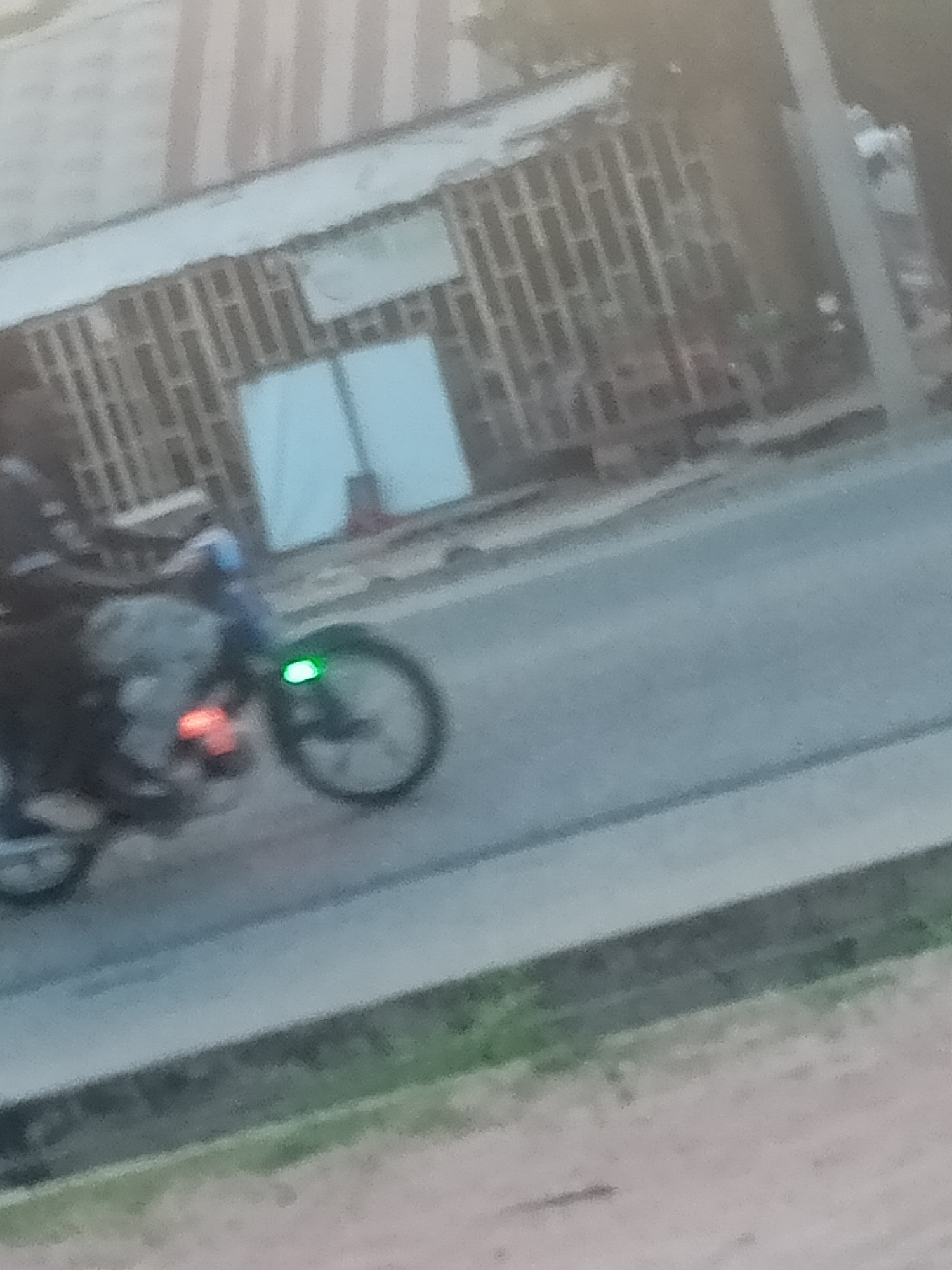 The image shows a street scene in Jalingo, Nigeria. A person is riding a motorcycle down a paved road. The motorcycle has a bright green light on its front wheel and a red light on its rear. The background consists of a building with a patterned facade, possibly a residential building or a shop. The overall image is blurry, suggesting it was taken from a moving vehicle or in low light conditions. The environment appears to be a typical urban street in Jalingo, with a sidewalk and some vegetation visible.