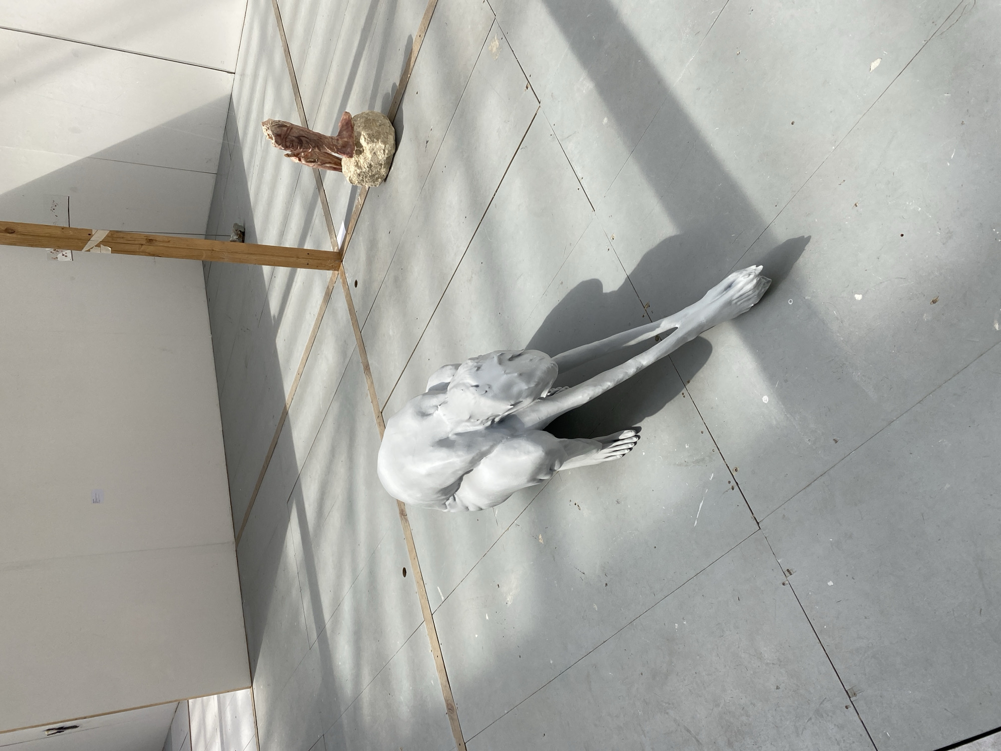 The scene is an art studio in Paris, France. Two sculptures are present on a floor consisting of large gray tiles. One sculpture is a life-size, pale gray figure of a person seated with their legs extended and arms folded in front. The other sculpture, in the background, is a brown and white organic form set atop a wooden frame. The lighting is bright and natural, creating sharp shadows. The walls are white and made of panels.